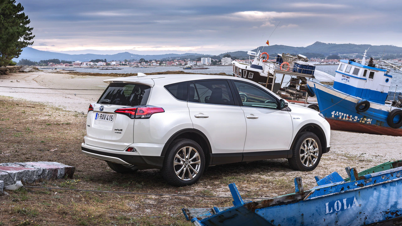 Weißer SUV parkt am Strand neben Fischerbooten bei bewölktem Himmel, mit Hintergrund von Hügeln und einer Stadt.