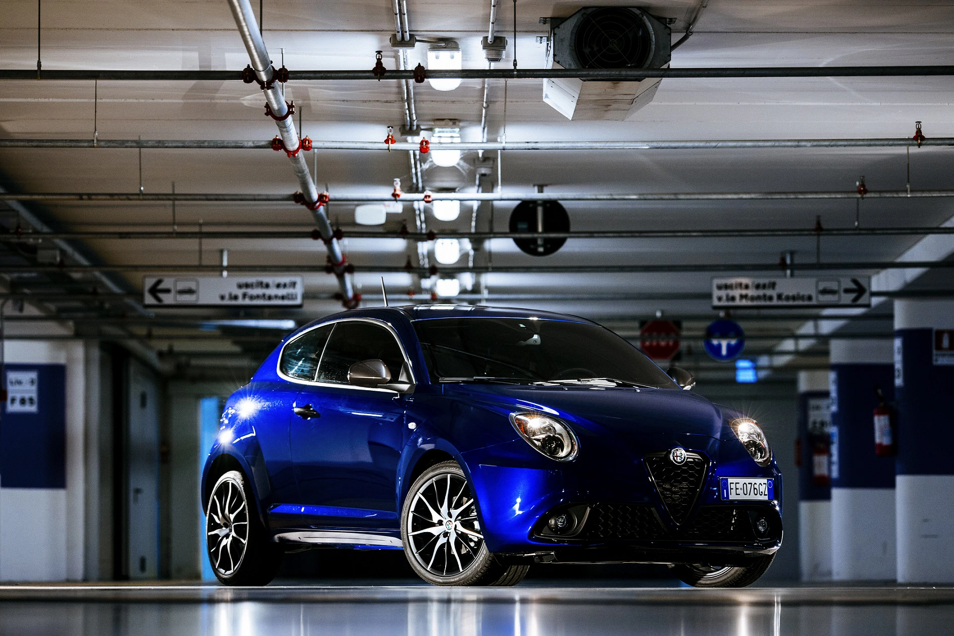 Blauer Alfa Romeo im beleuchteten Parkhaus, umgeben von weißen Säulen und Wegweisern. Dynamische Beleuchtung betont das sportliche Design.