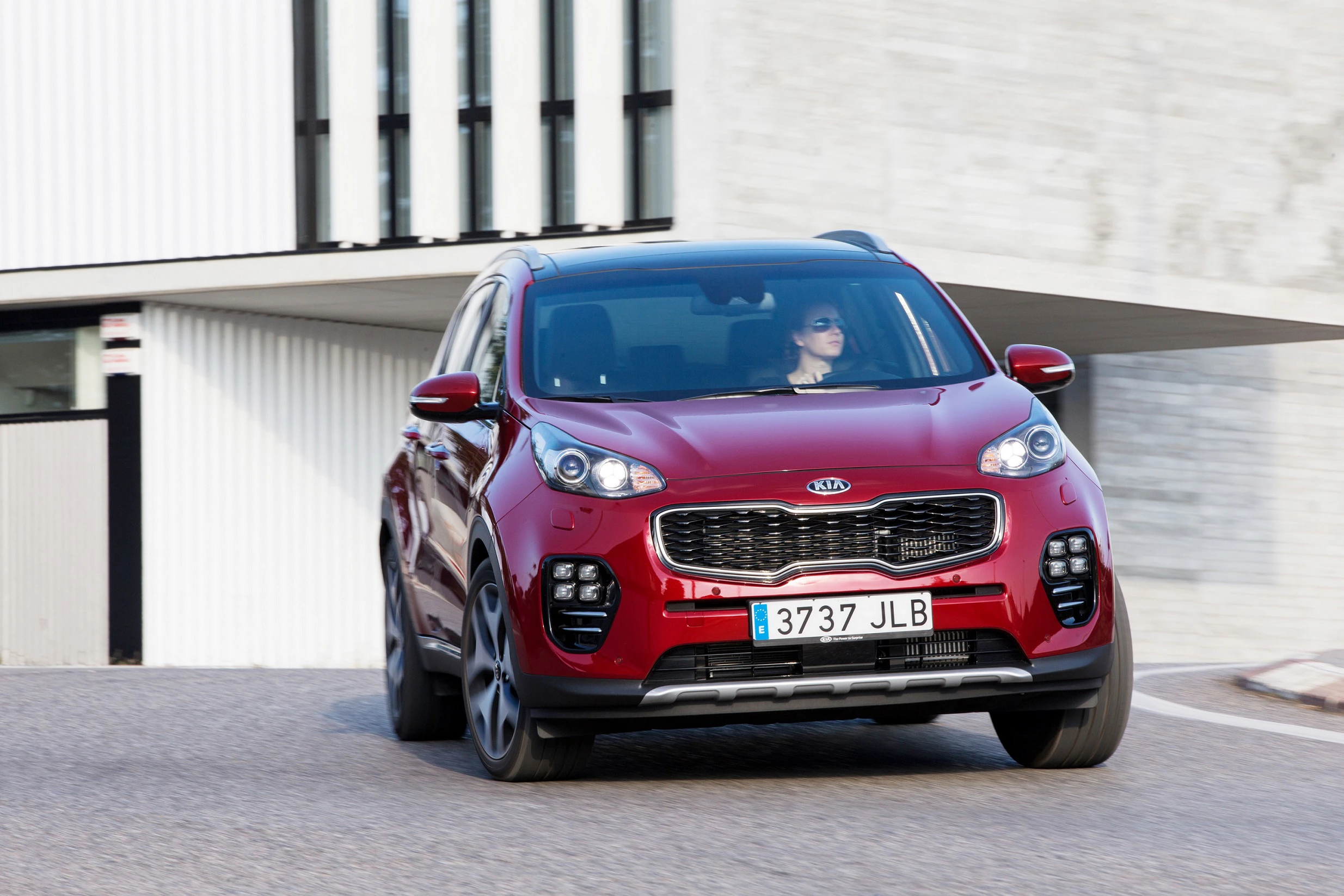 Roter SUV fährt eine Kurve entlang einer modernen Gebäudefassade. Eine Person sitzt am Steuer. Autokennzeichen: 3737 JLB.