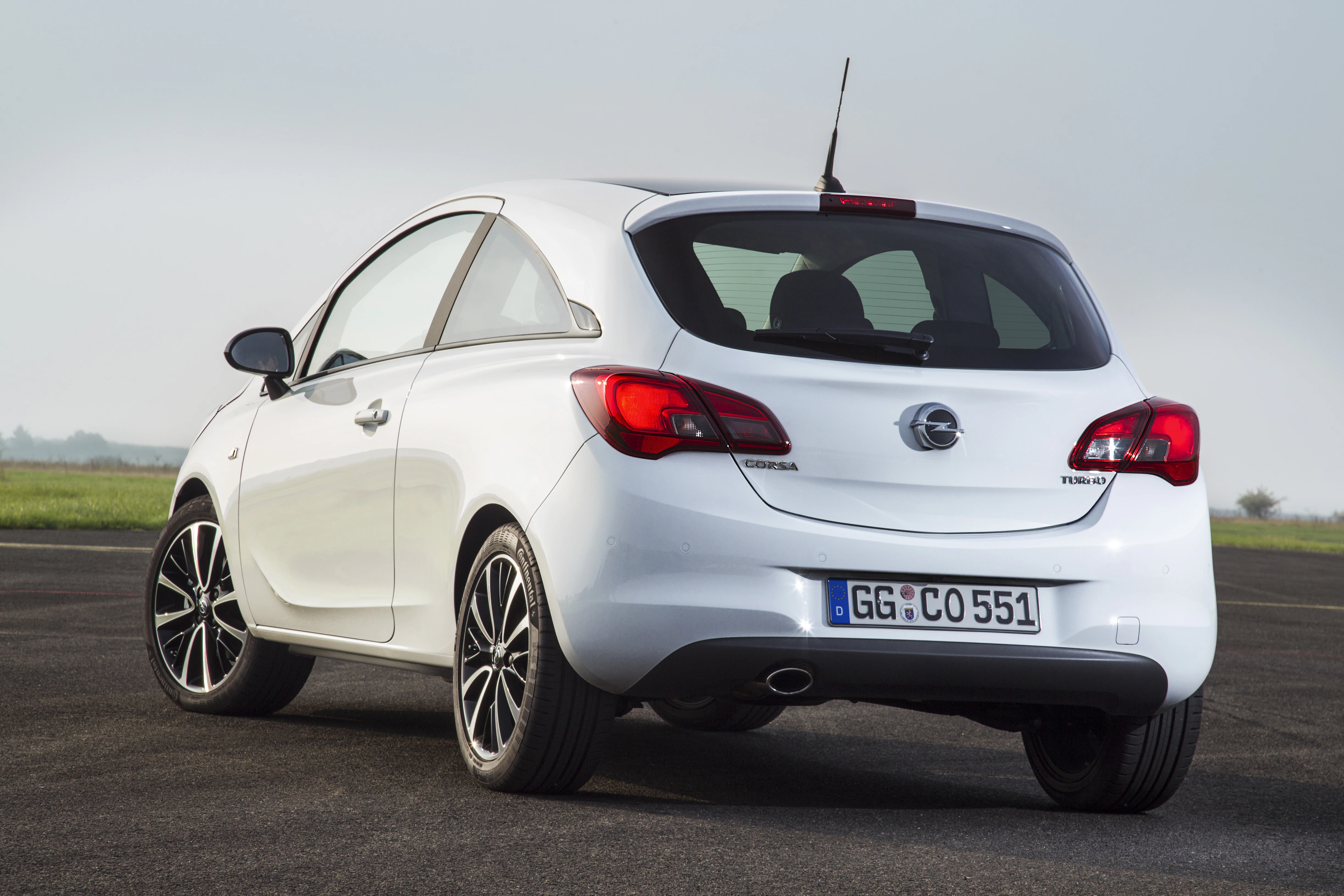 Weißer Opel Corsa auf einer asphaltierten Fläche, Rückansicht. Wetter klar, Horizont im Hintergrund sichtbar.