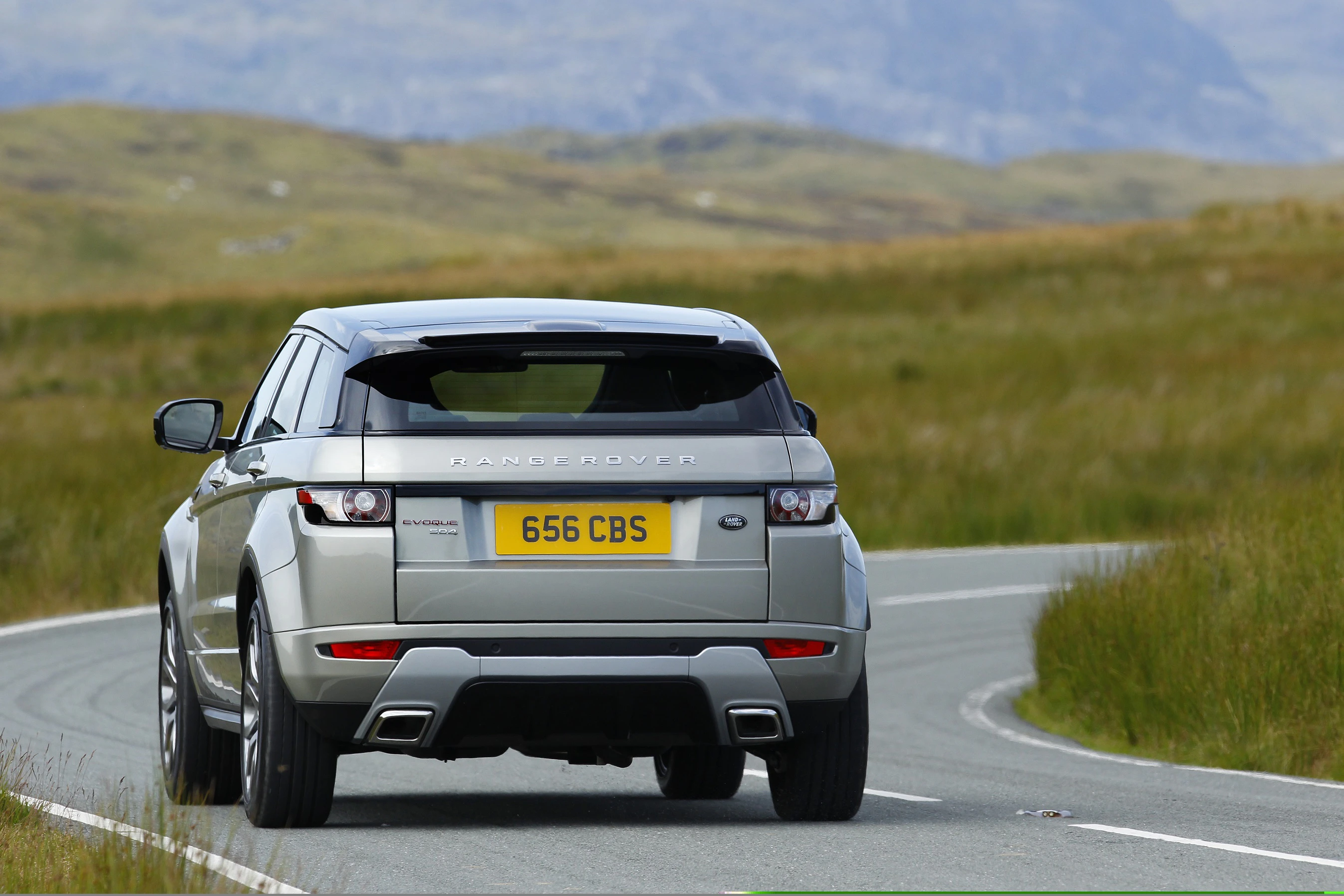 Silberner Range Rover fährt auf einer kurvenreichen Straße durch eine grüne, hügelige Landschaft.