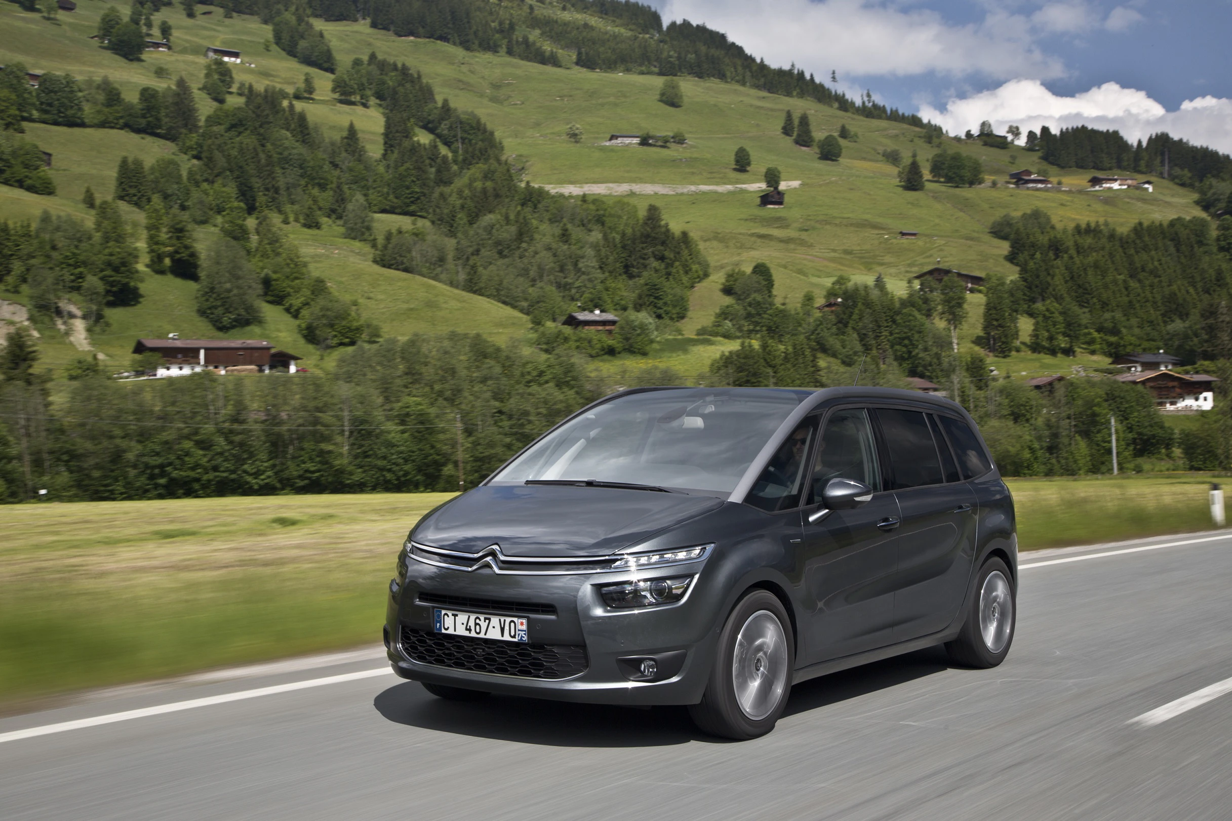 Ein grauer Citroën C4 Picasso fährt auf einer Landstraße durch eine idyllische, grüne Hügellandschaft mit vereinzelten Holzhäusern.