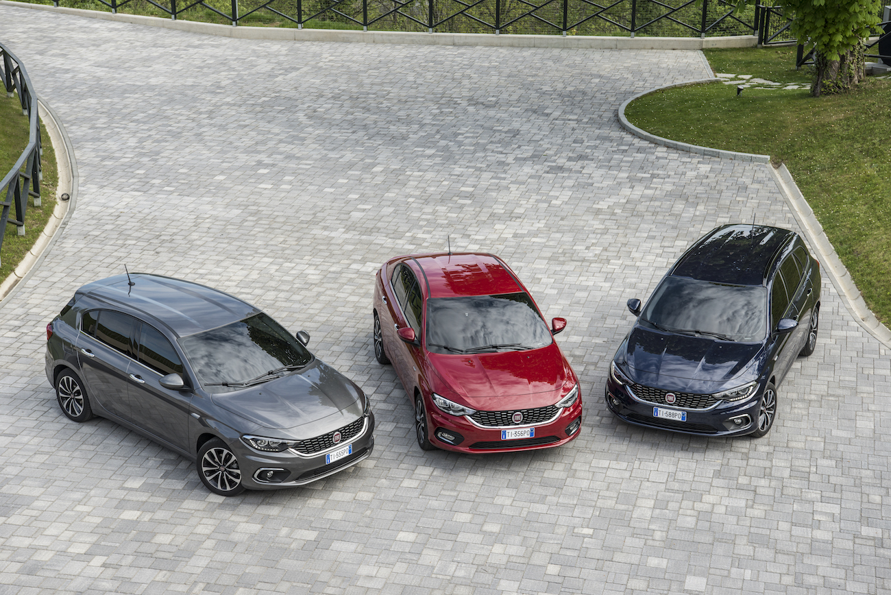 Drei Autos in verschiedenen Farben (Grau, Rot, Schwarz) stehen nebeneinander auf einem gepflasterten Parkplatz mit grünen Bäumen im Hintergrund.