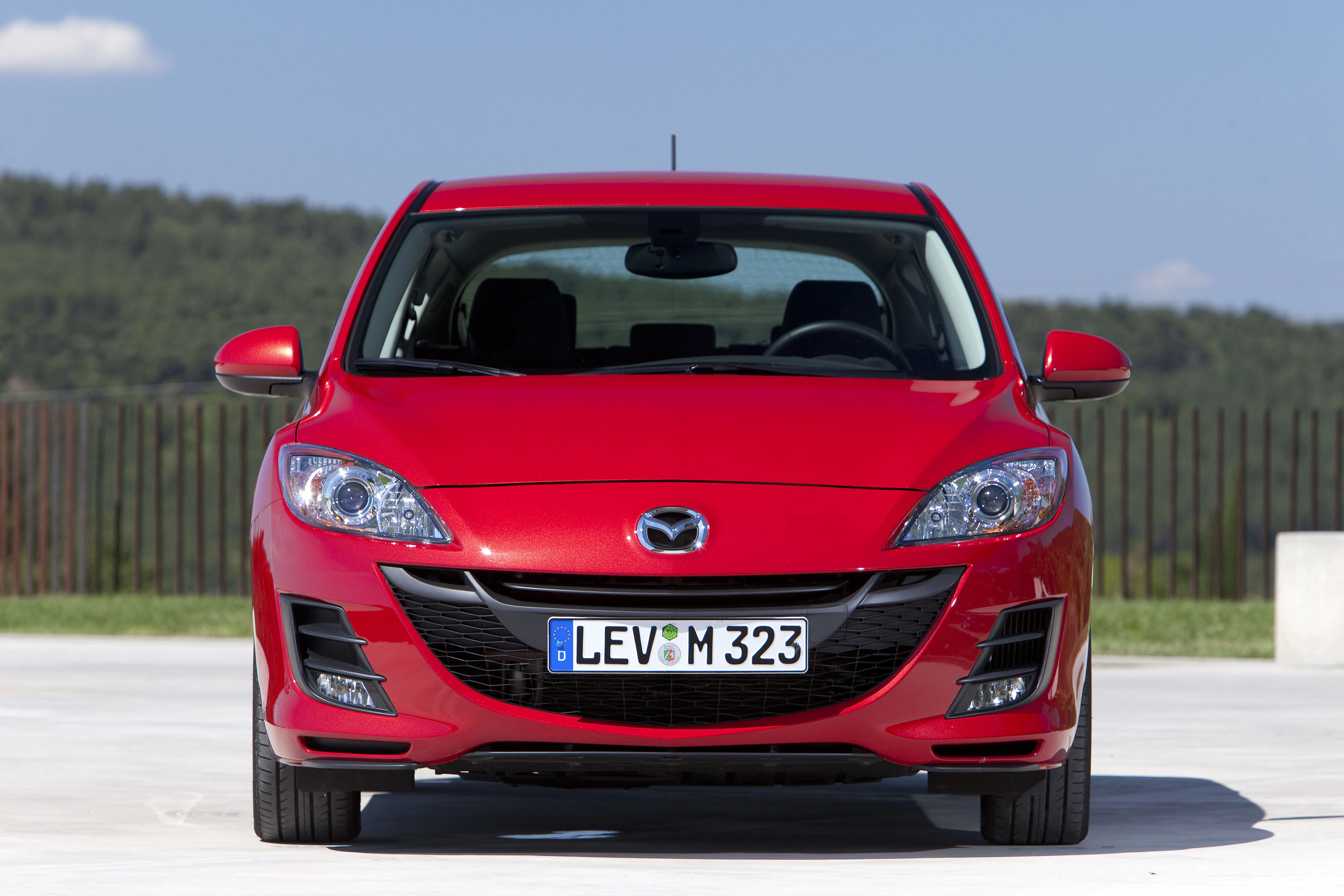 Roter Mazda mit deutscher Kennzeichen, frontalansicht, auf Parkplatz mit bewaldetem Hintergrund unter blauem Himmel.