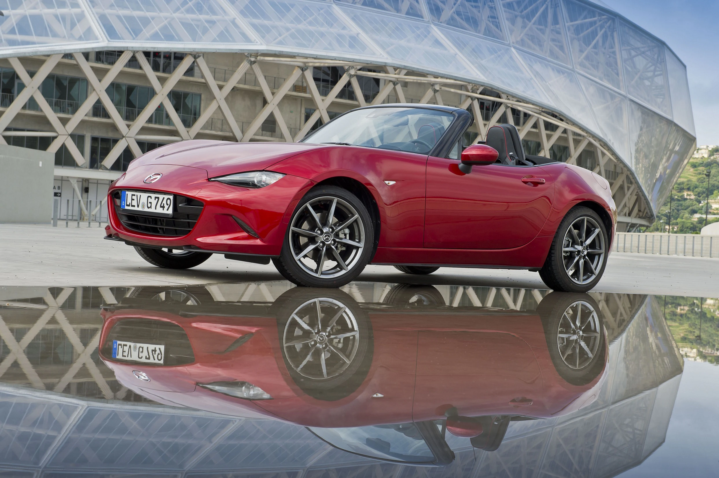 Roter Sportwagen vor moderner Architektur, spiegelt sich auf nasser Oberfläche wider. Klarer Himmel und geometrisches Design im Hintergrund.