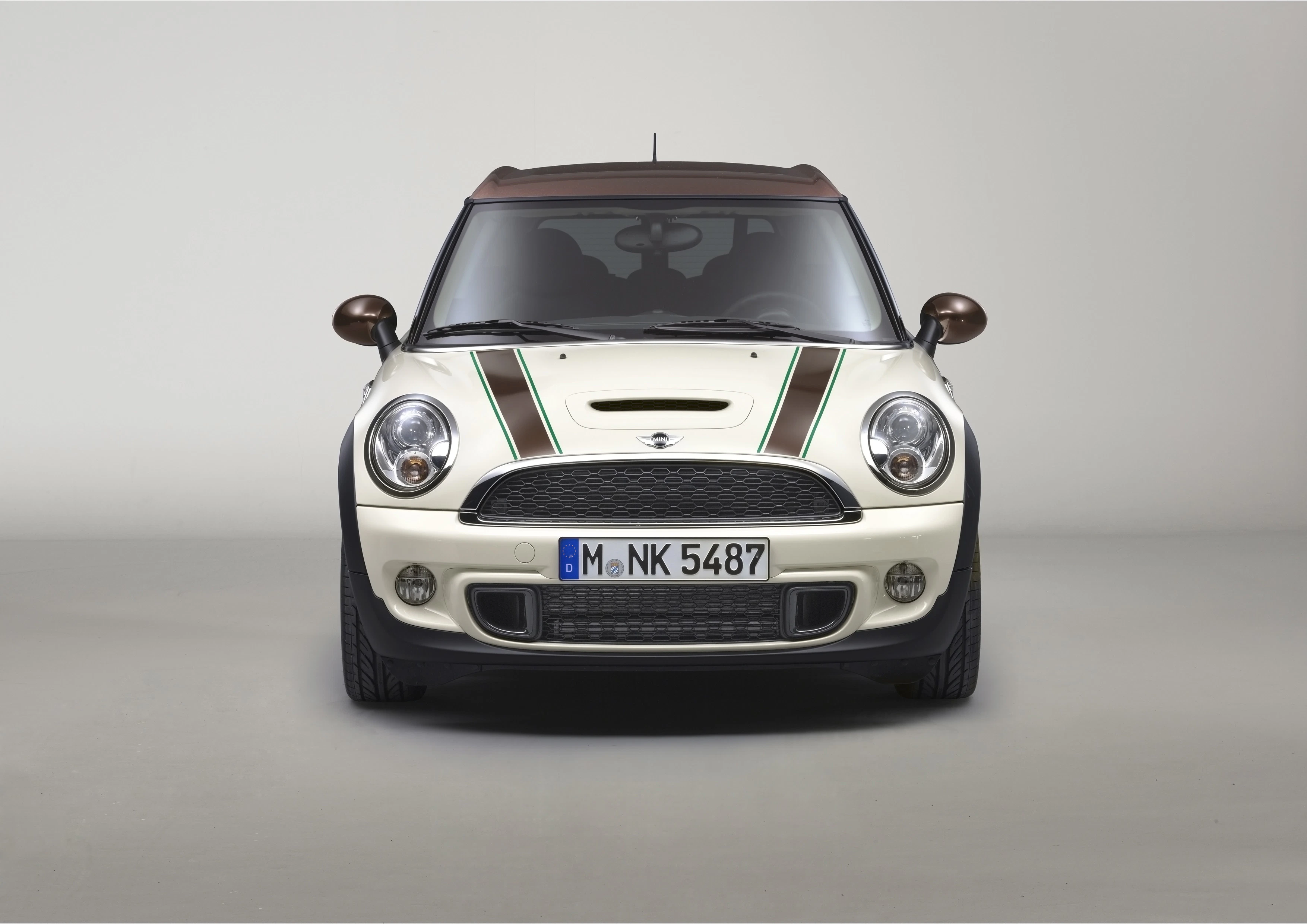 Weißer Mini Cooper in Frontalansicht mit grünen Streifen auf der Motorhaube und braunen Seitenspiegeln, vor neutralem Hintergrund.