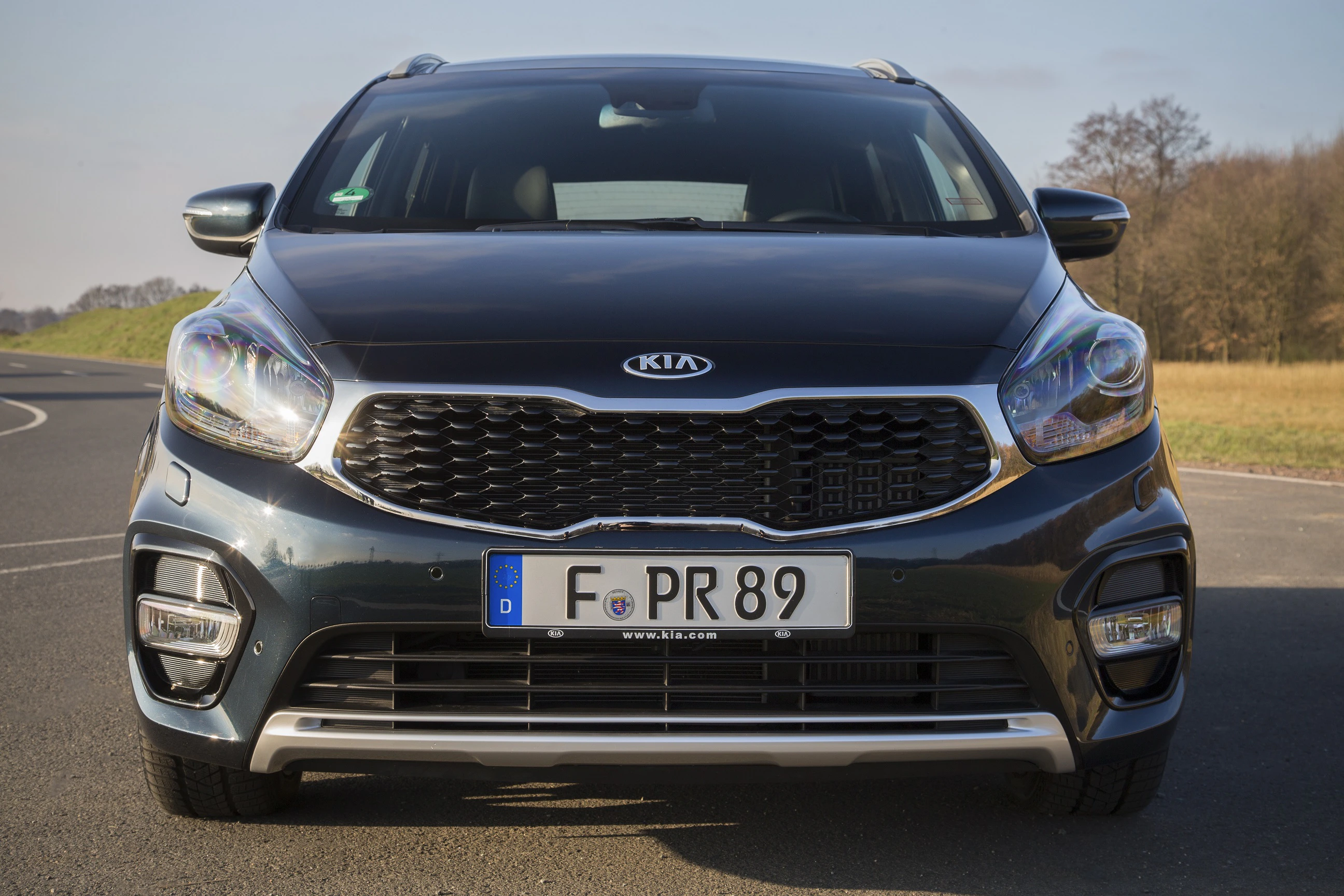 Frontansicht eines Kia-Autos auf einer Landstraße bei klarem Himmel, deutsche Nummernschilder sichtbar.