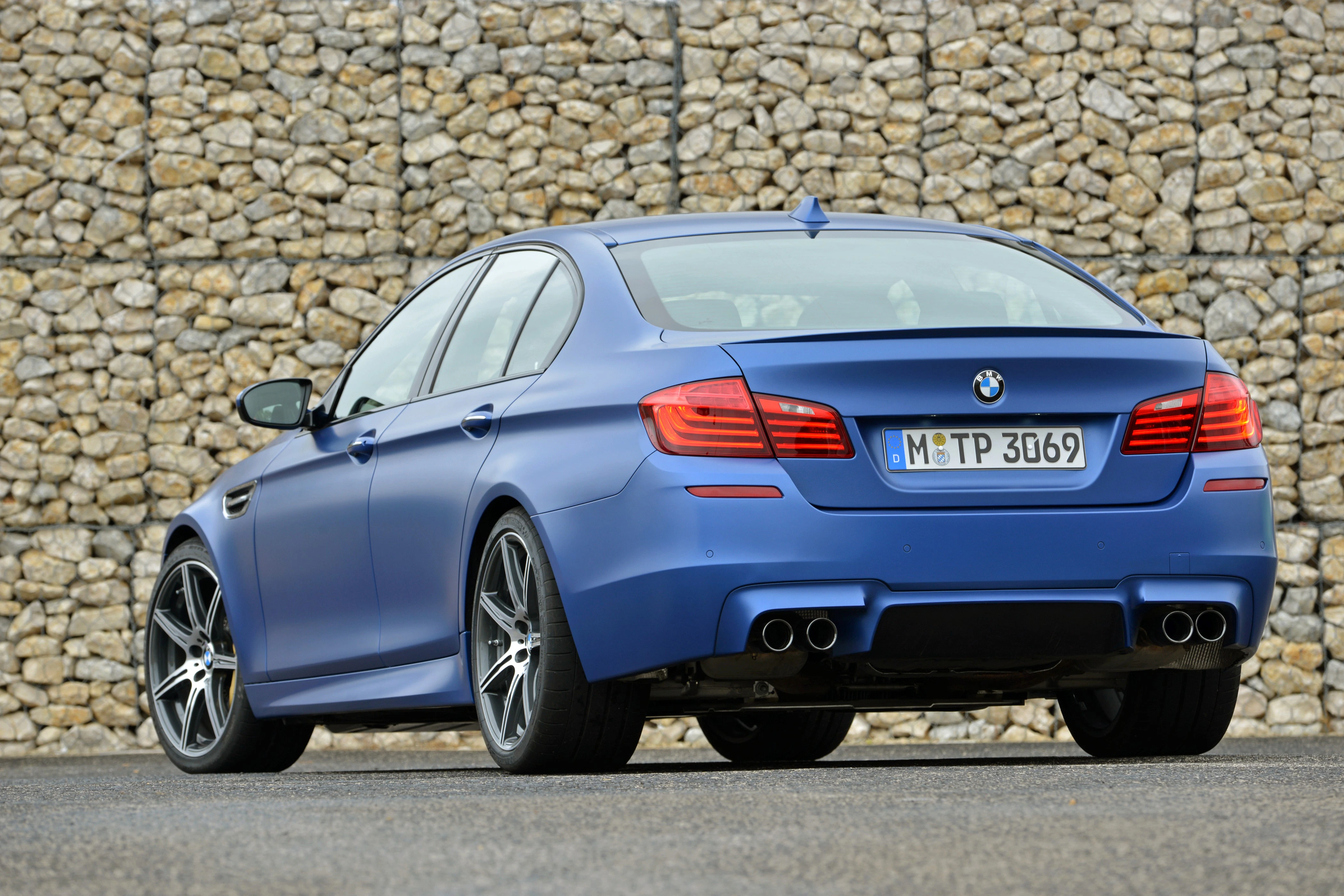 Blauer BMW auf Asphalt vor einer Steinwand, Heckansicht. Der Wagen hat sportliche Auspuffrohre und moderne Felgen.