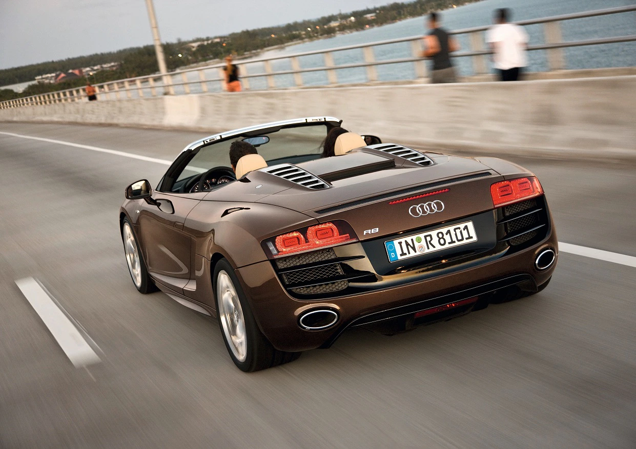 Brauner Audi R8 Cabrio fährt über eine Brücke am Wasser entlang, bei leicht bedecktem Himmel. Heckansicht des Sportwagens.
