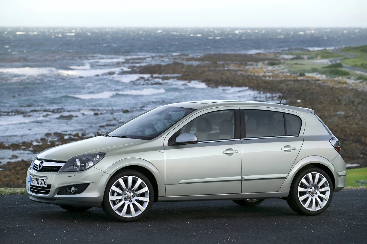 Silberner Opel Astra steht auf einer Küstenstraße, im Hintergrund das Meer und felsige Küste an einem bewölkten Tag.