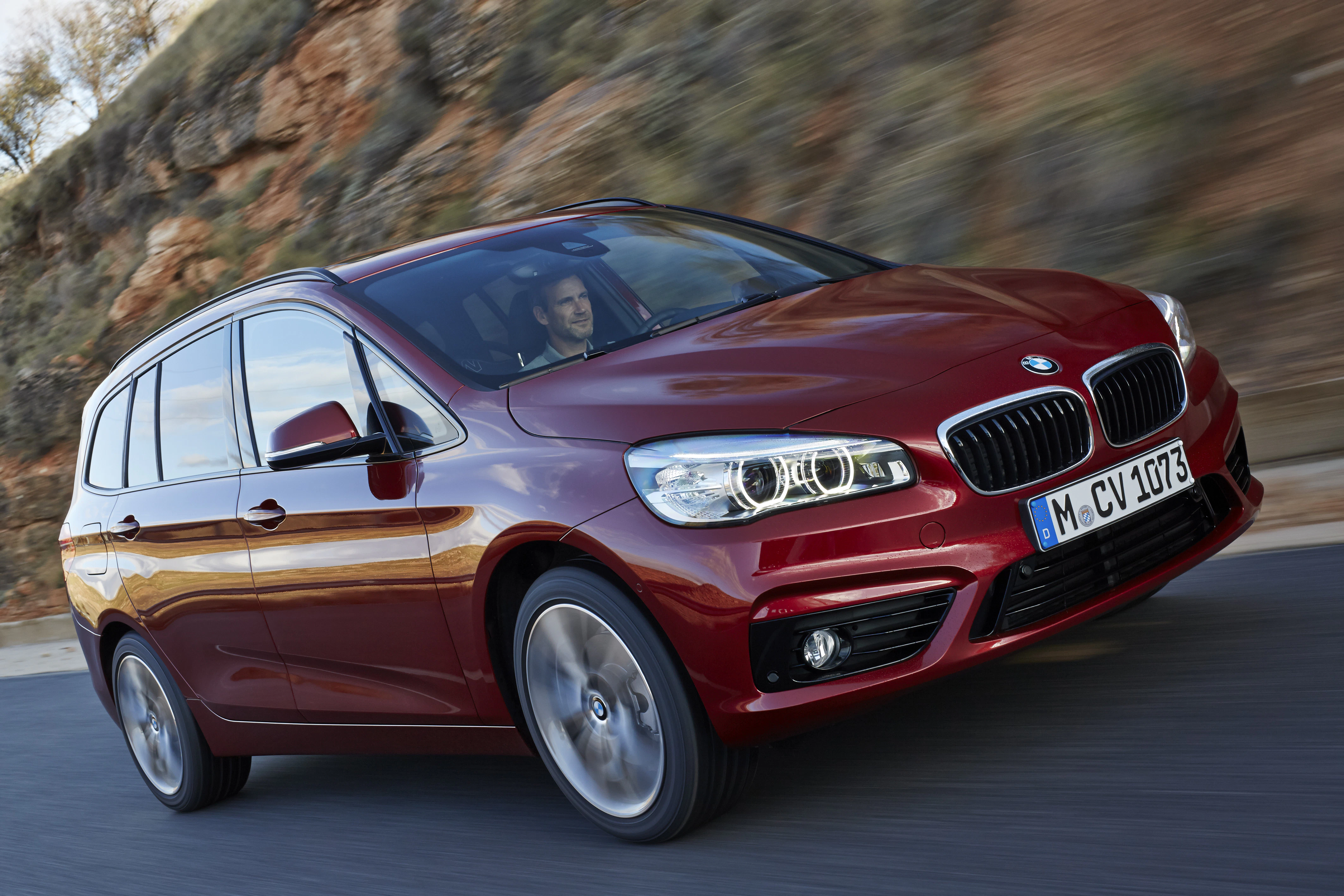 Roter BMW fährt dynamisch auf einer Landstraße, seitliche Frontansicht, Fahrer sichtbar. Hintergrund: Felsen und Bäume.
