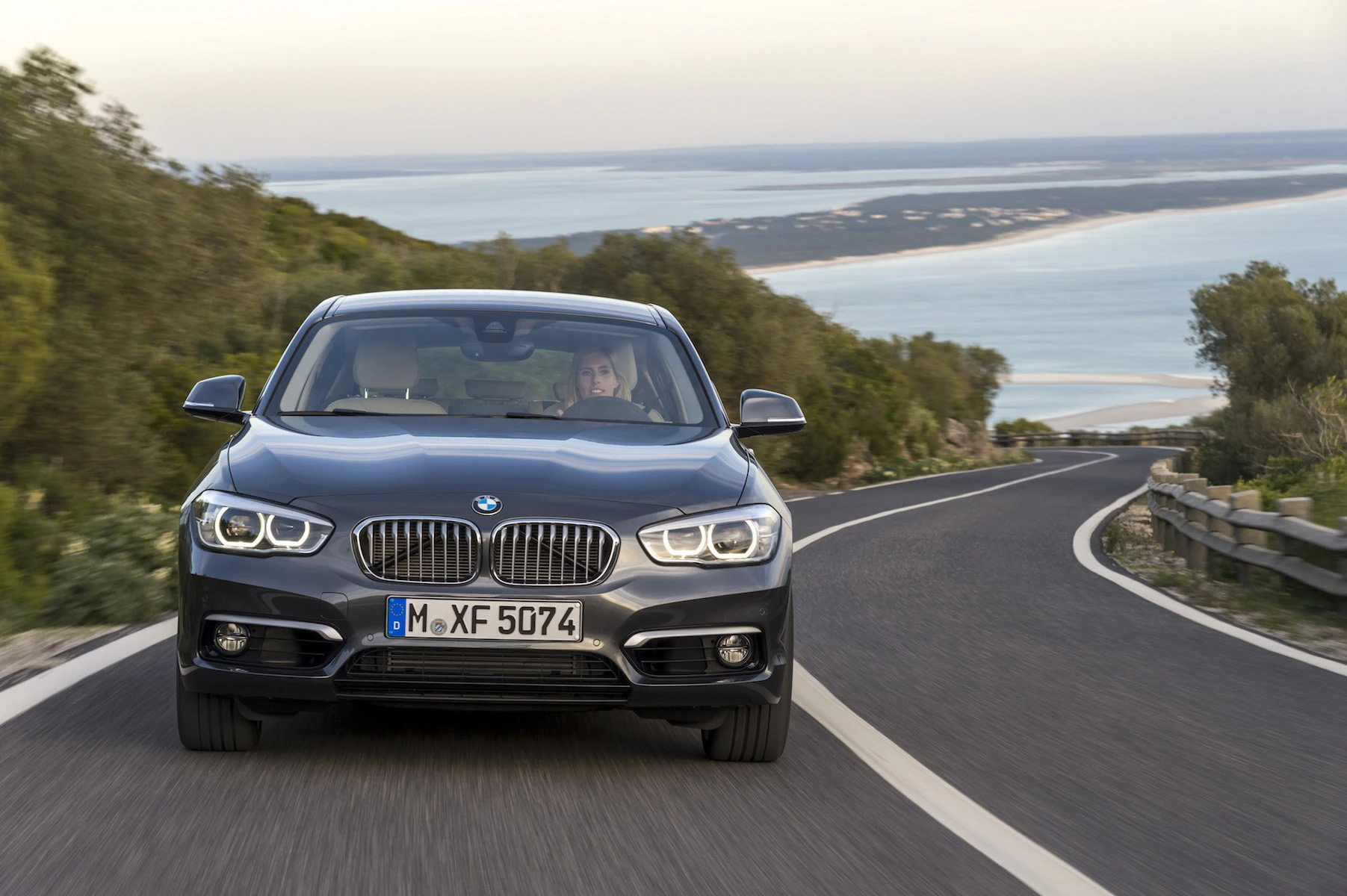 Ein grauer BMW fährt auf einer kurvigen Straße am Meer entlang, umgeben von Bäumen und blauem Himmel im Hintergrund.
