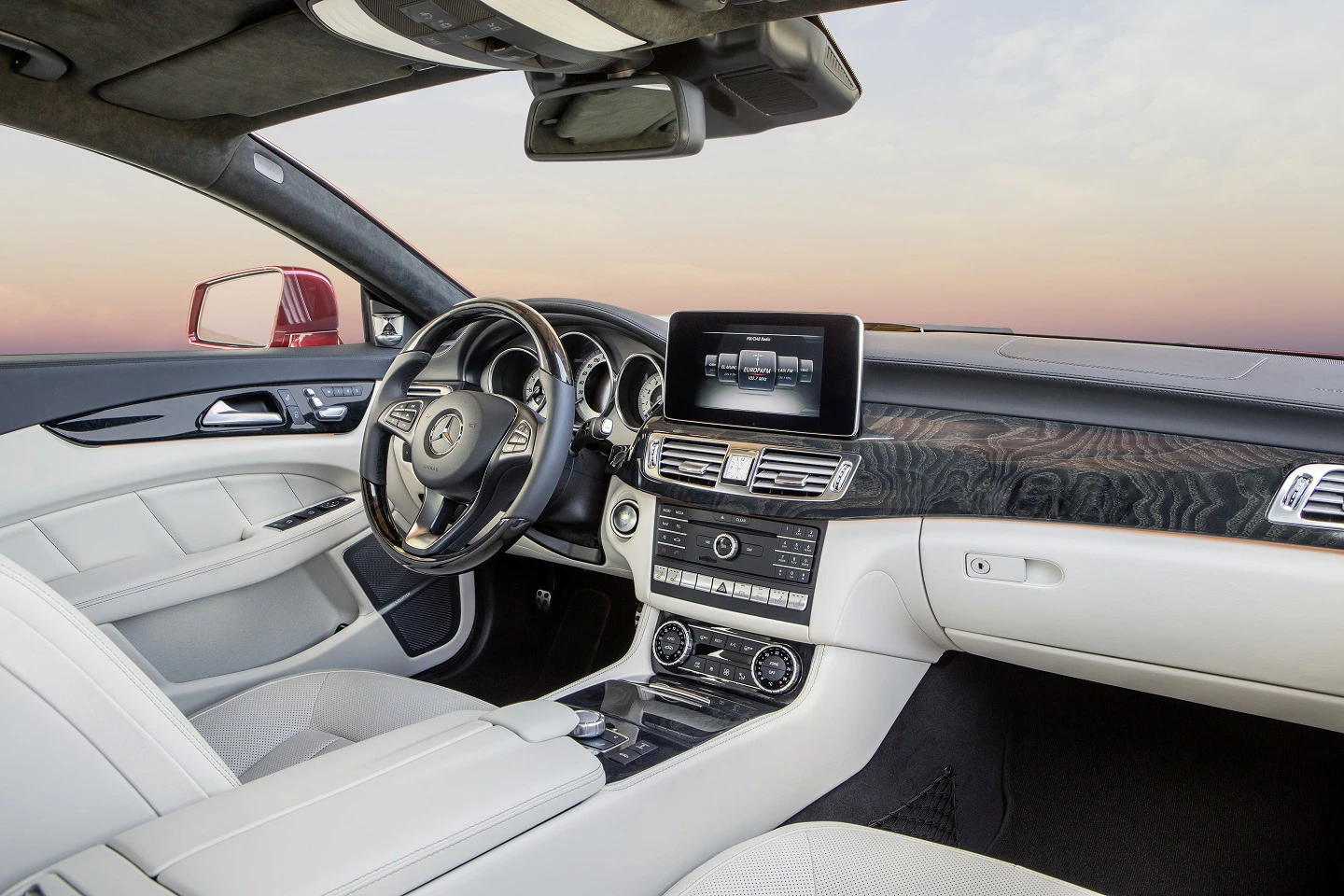 Innenraum eines luxuriösen Autos mit hellen Ledersitzen, modernem Armaturenbrett, integriertem Bildschirm und Holzverkleidung, bei Sonnenuntergang.