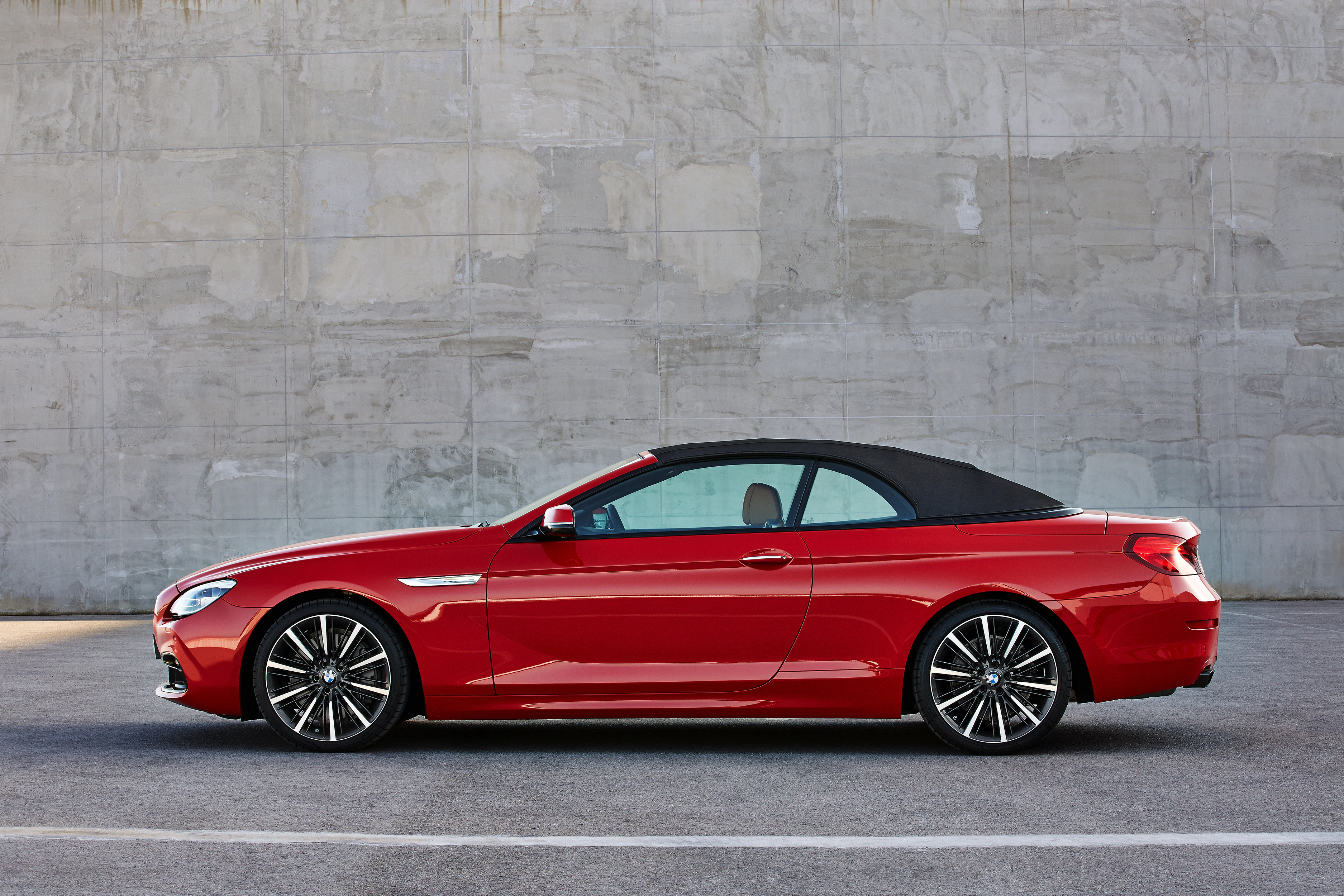 Rotes Cabriolet mit schwarzem Verdeck, seitlich vor einer grauen Betonwand geparkt.