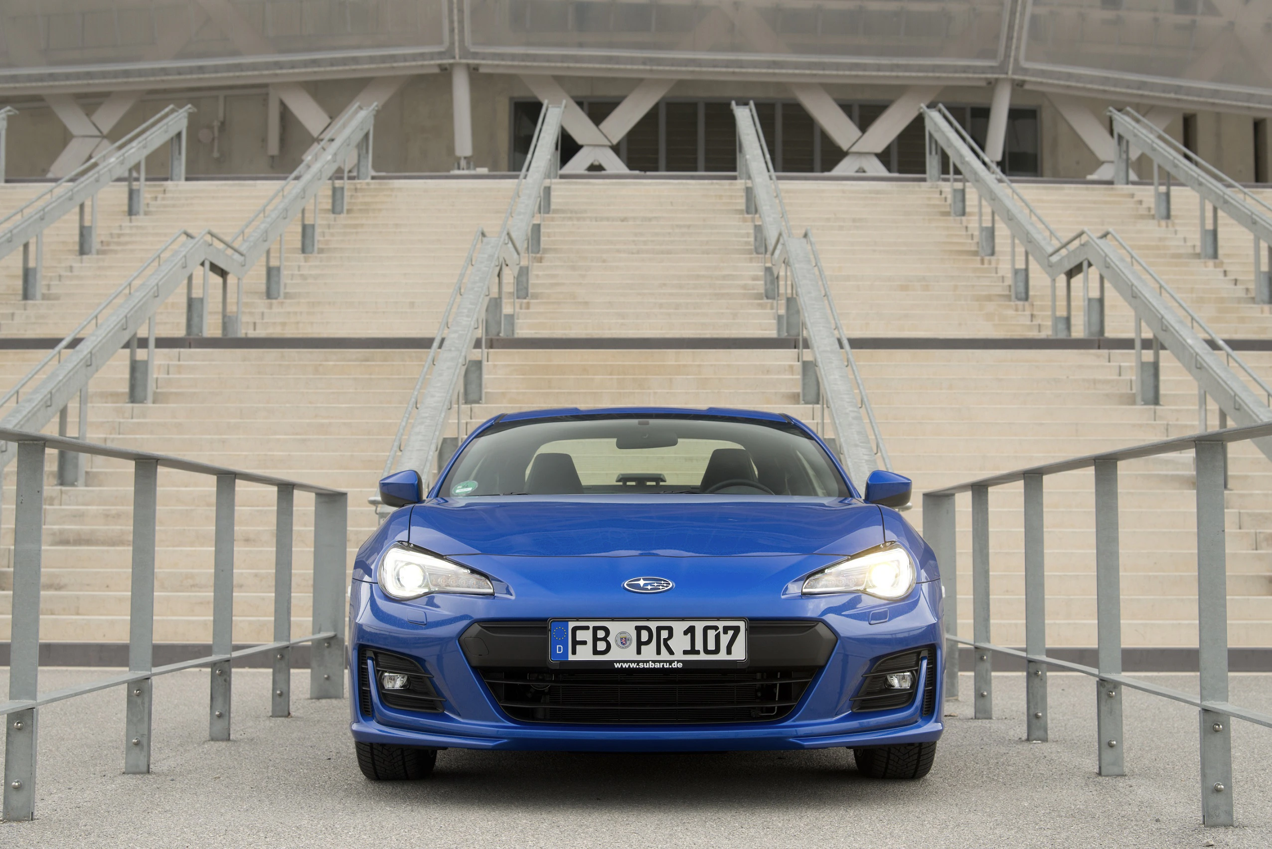 Blauer Sportwagen auf einem Parkplatz vor einer breiten Treppe. Im Hintergrund modernes Gebäude. Nr. Schild: FB-PR 107.
