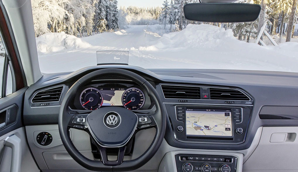 Blick aus dem Innenraum eines VW auf schneebedeckte Straße, Lenkrad im Vordergrund, Infotainmentsystem zeigt Karte an, verschneiter Wald im Hintergrund.