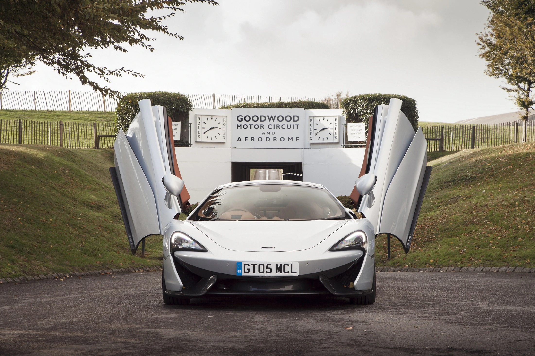 Ein silberner Sportwagen mit geöffneten Flügeltüren vor dem Goodwood Motor Circuit Schild.