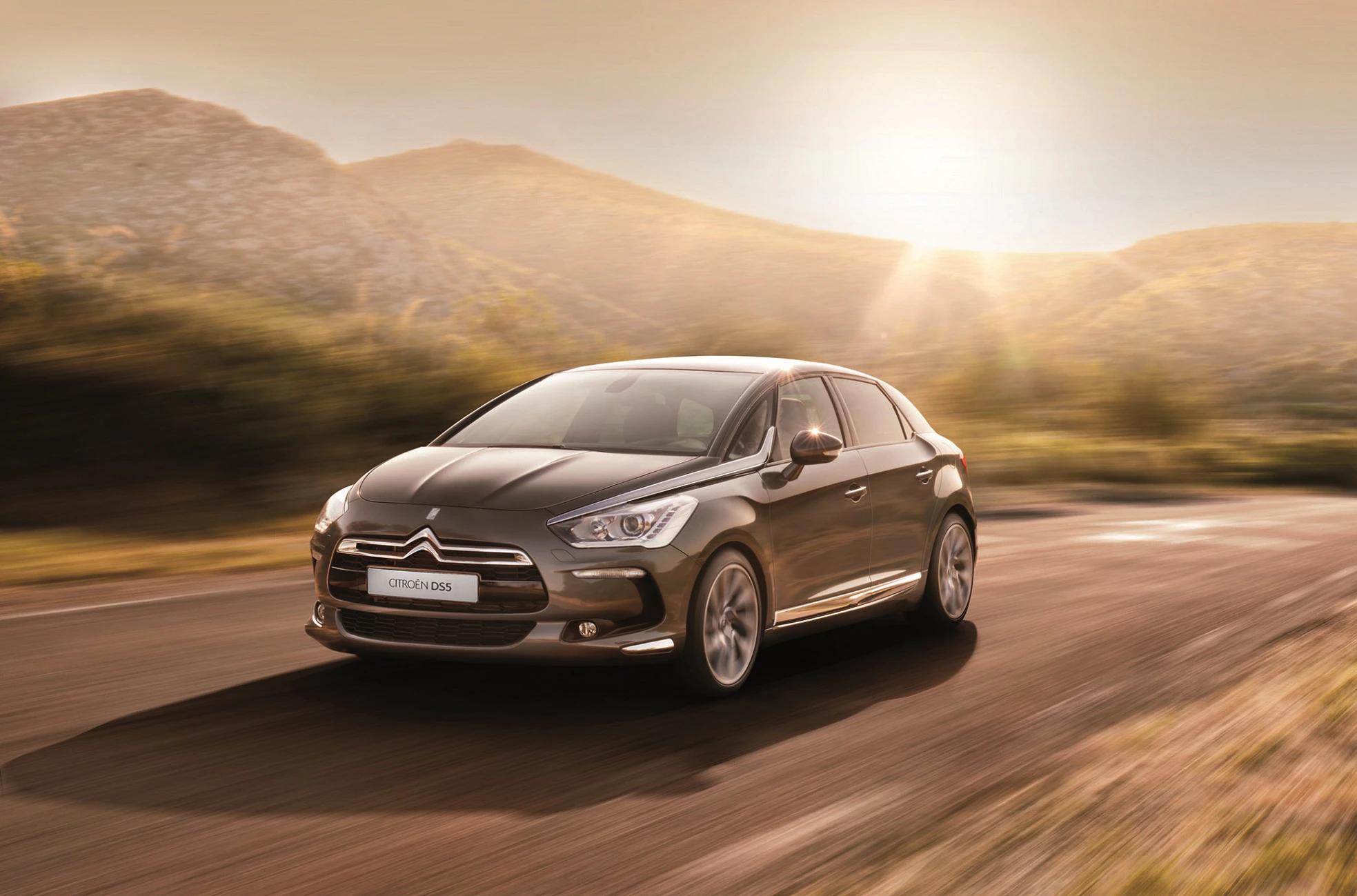 Ein dunkelgrauer Citroën DS5 fährt auf einer malerischen Landstraße bei Sonnenuntergang, umgeben von Bergen und goldener Abendstimmung.