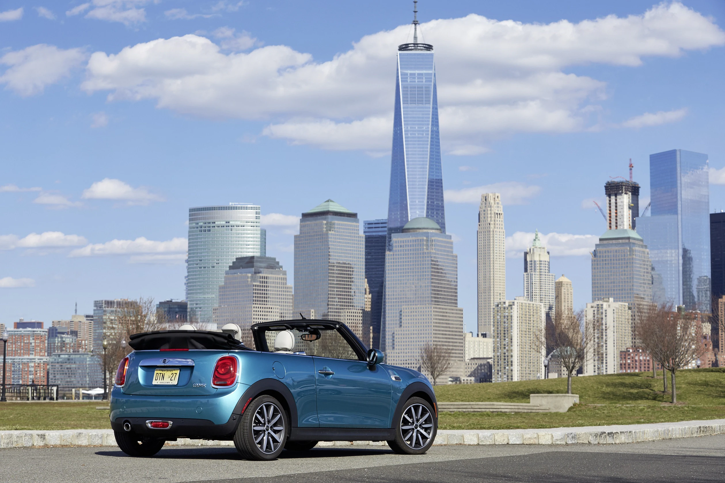 Blauer Mini Cooper Cabrio vor der Skyline von New York City mit dem One World Trade Center im Hintergrund und teilweise bewölktem Himmel.