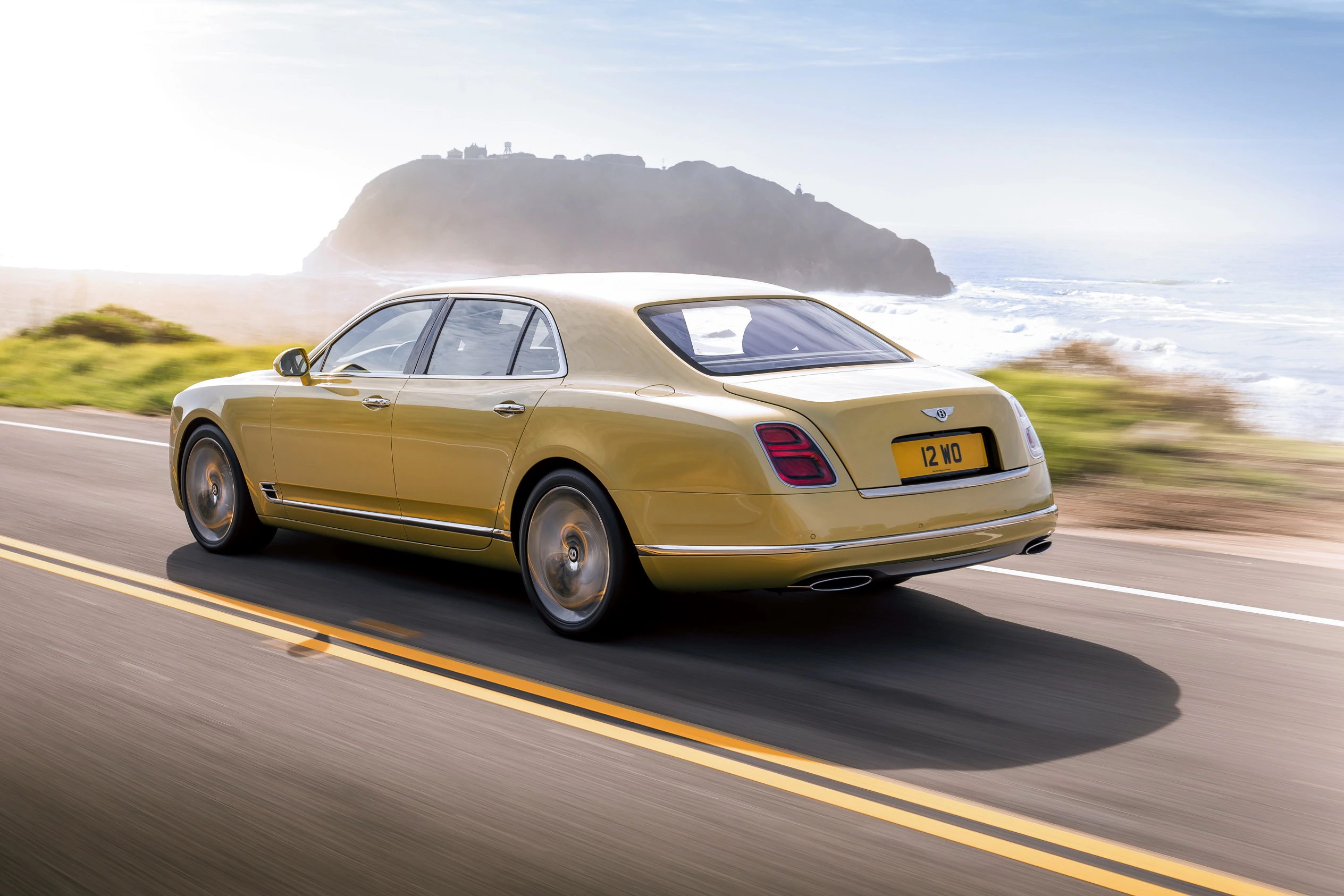 Luxuriöser, goldfarbener Bentley fährt auf einer kurvigen Küstenstraße bei Sonnenschein. Im Hintergrund ein Hügel und das Meer.