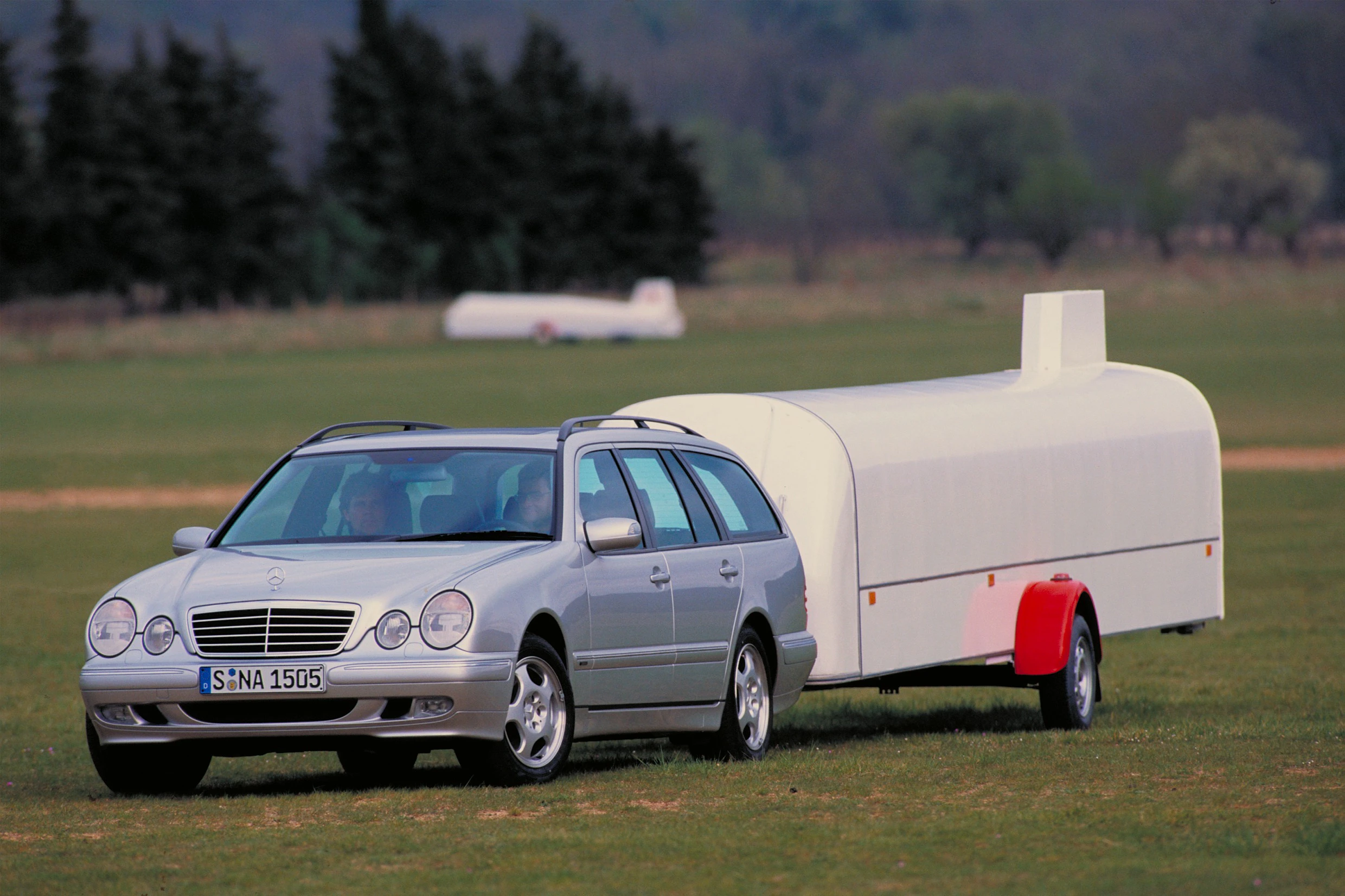Silberner Mercedes-Benz Kombi mit modernem, weißem Wohnwagenanhänger auf Wiese. Im Hintergrund Bäume und Feld.