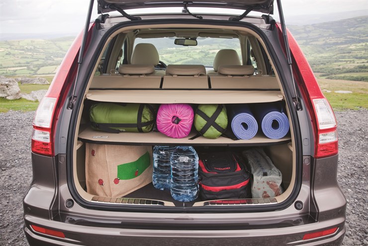 Kofferraum eines Autos mit Campingausrüstung: Schlafsäcke, Isomatten, eine Tasche, Wasserflaschen und andere Utensilien vor ländlicher Landschaft.