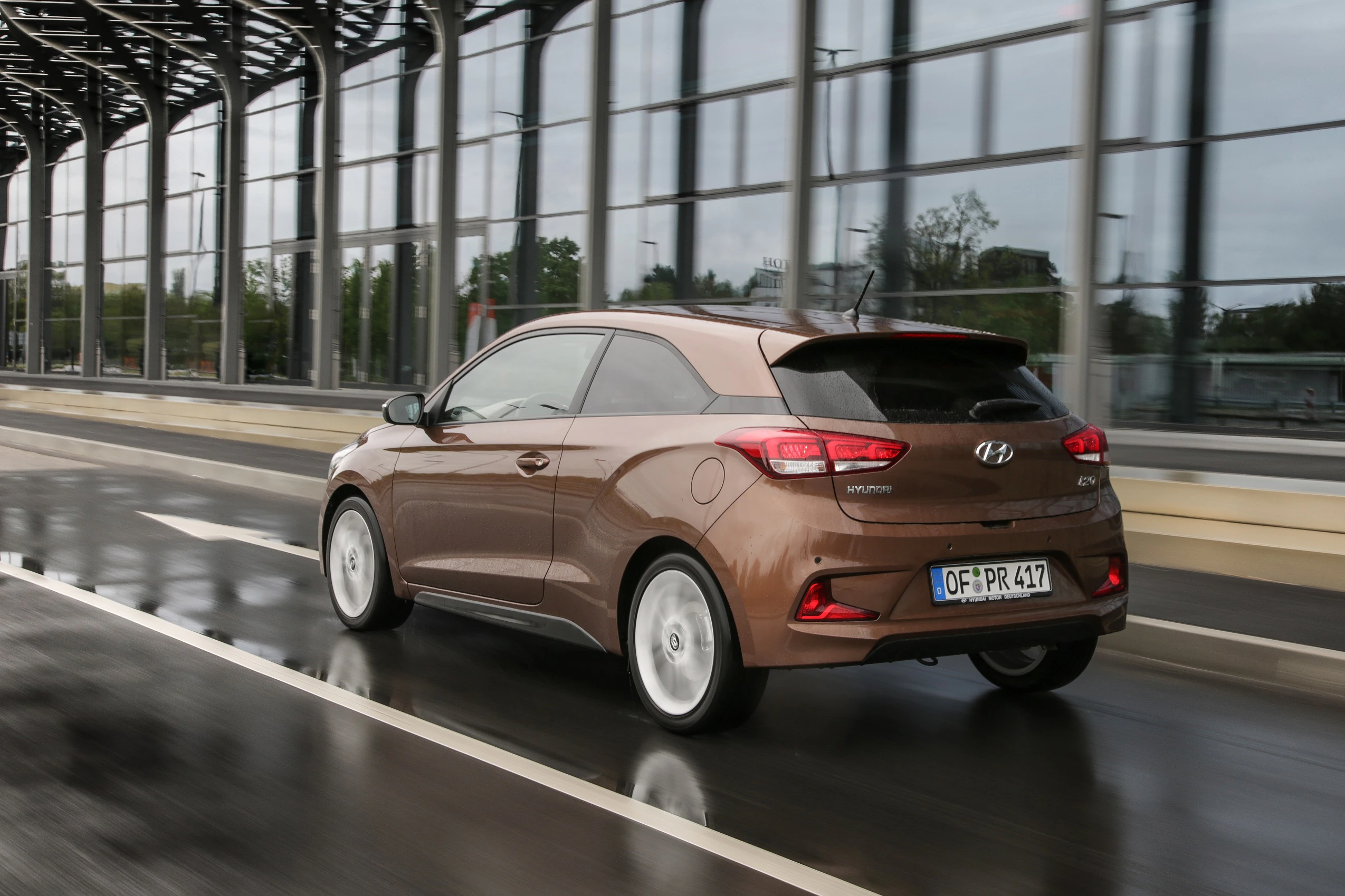 Brauner Hyundai i20 fährt auf regennasser Straße an einem modernen Gebäude vorbei.