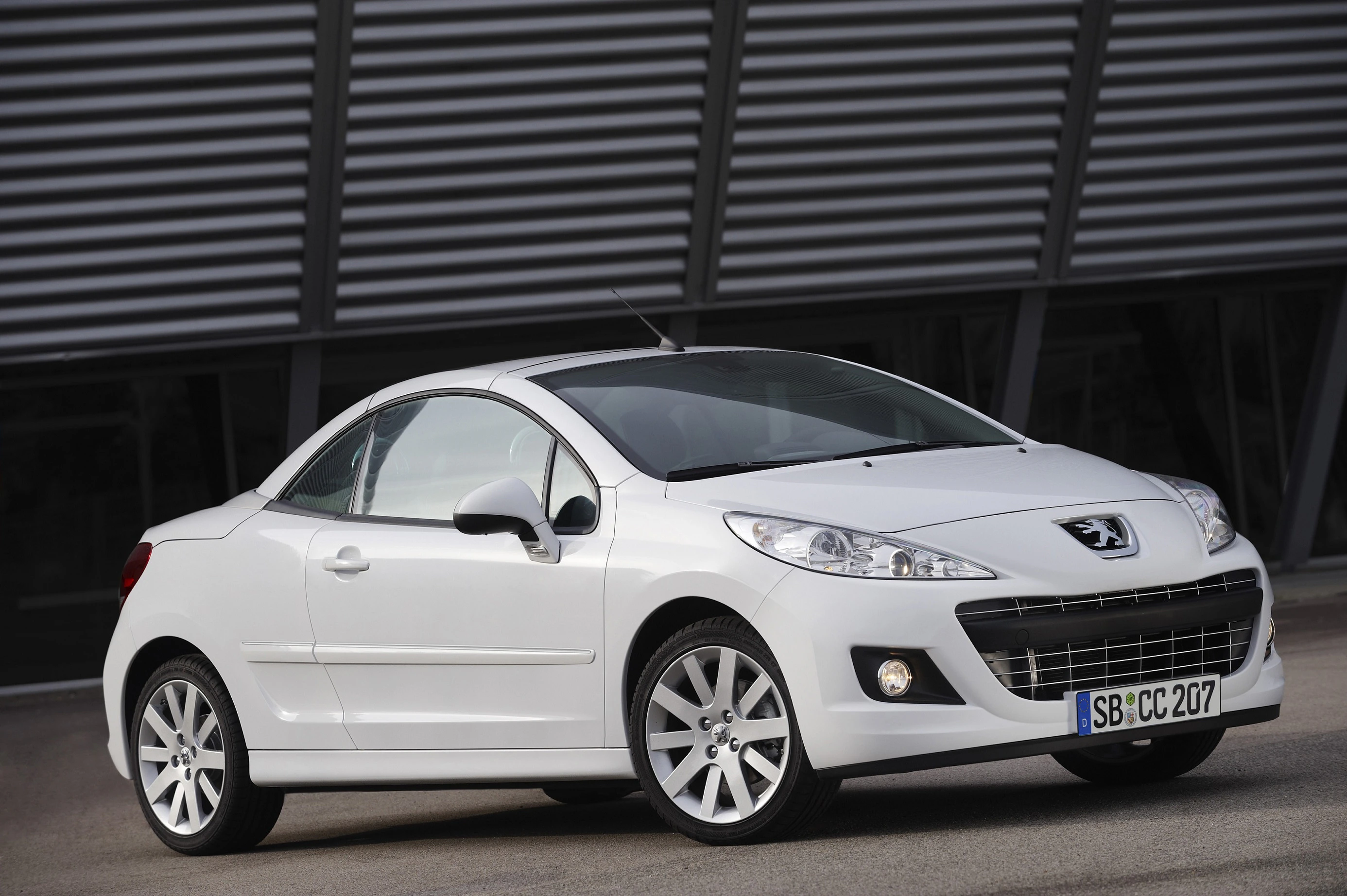 Weißer Peugeot 207 CC vor modernem Gebäude. Cabriolet mit geschlossenem Dach, Alufelgen und sportlichem Design.