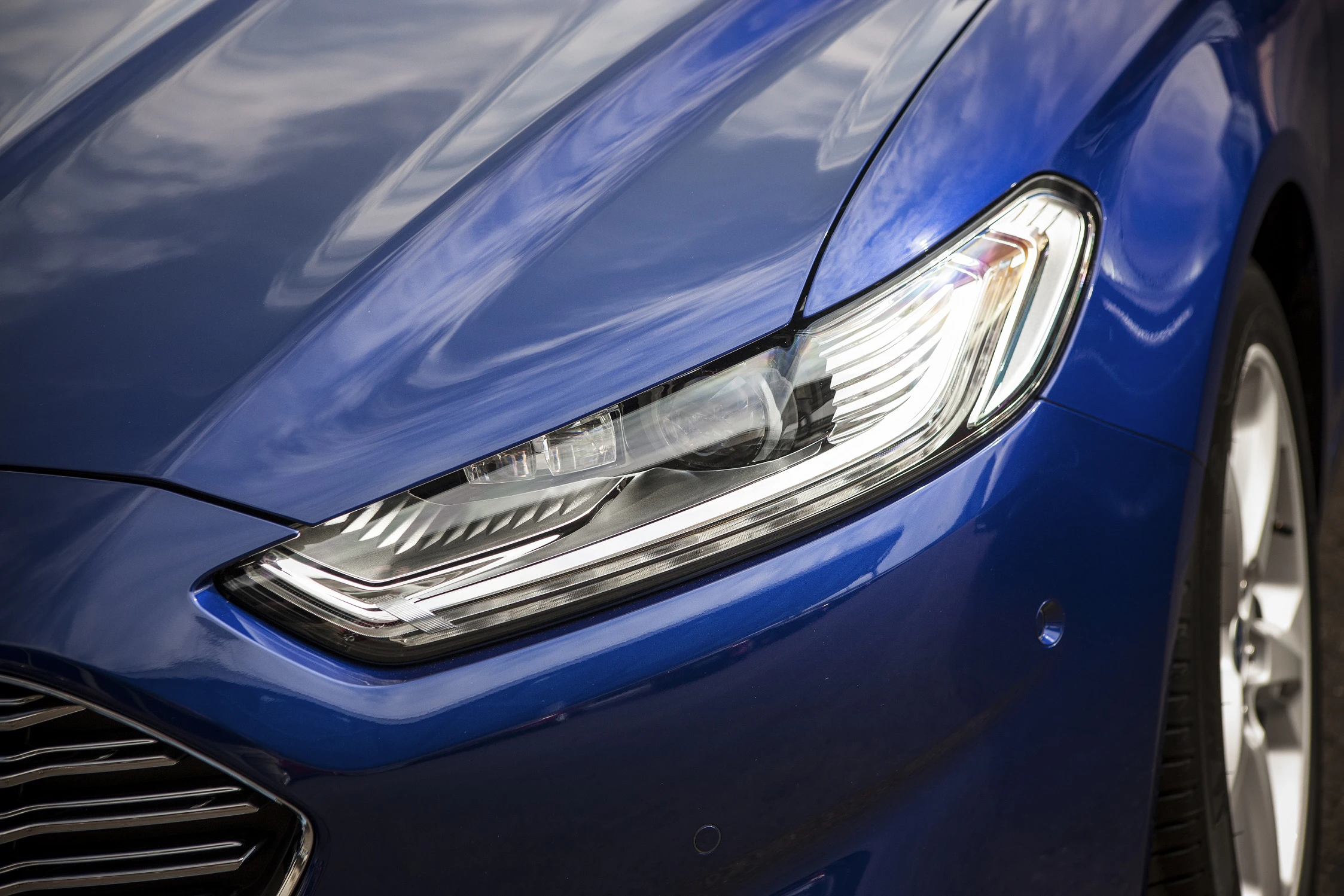 Nahaufnahme eines blauen Autos mit modernen Frontscheinwerfern und glänzender Lackierung, reflektierende Wolken im Hintergrund.