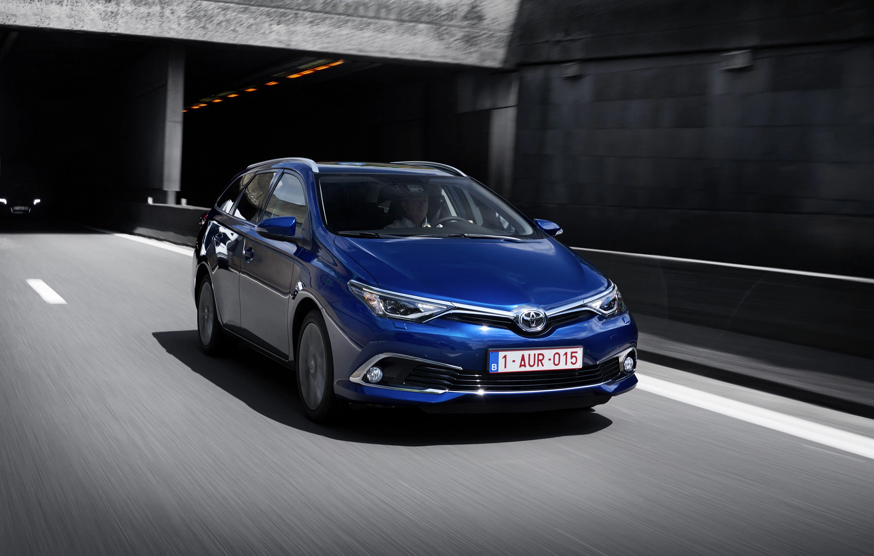 Blauer Toyota fährt durch einen Tunnel auf einer dreispurigen Straße bei Tageslicht. Frontansicht des Wagens, belgisches Nummernschild sichtbar.