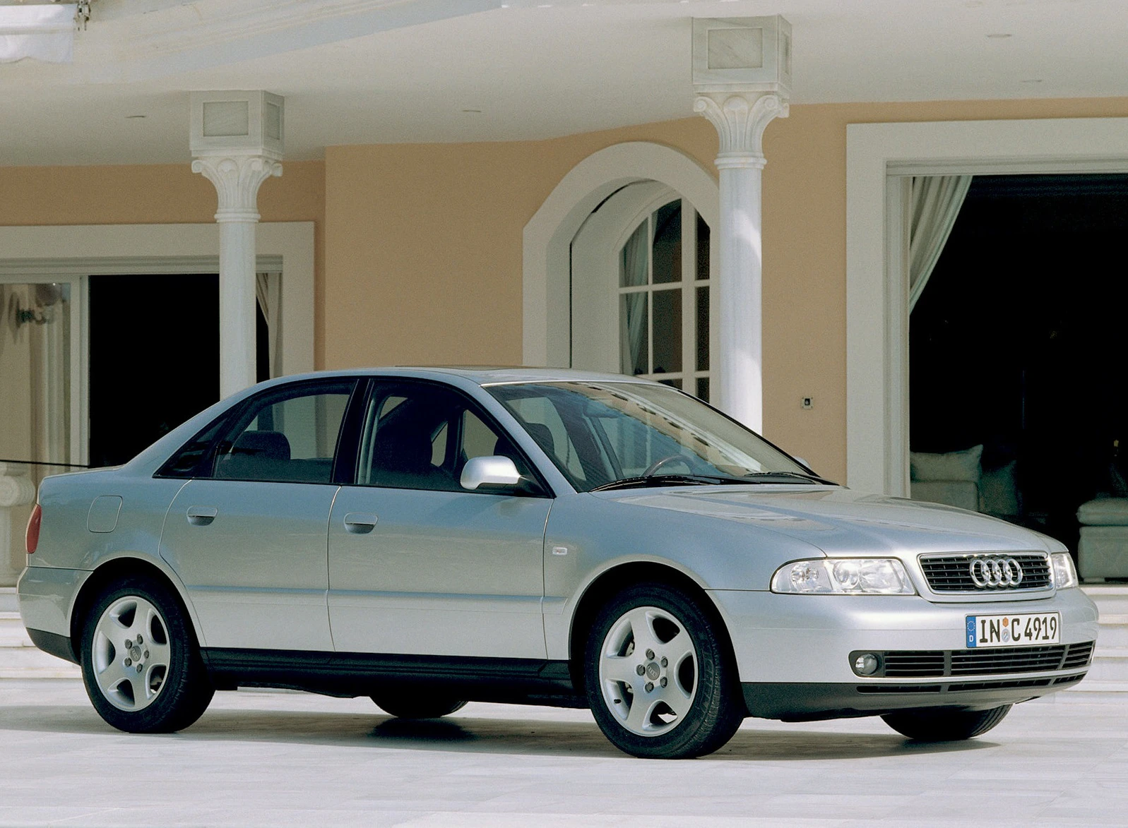 Silberner Audi Limousine der 1990er Jahre steht vor einem eleganten, hellen Gebäude mit Säulen und großen Fenstern.