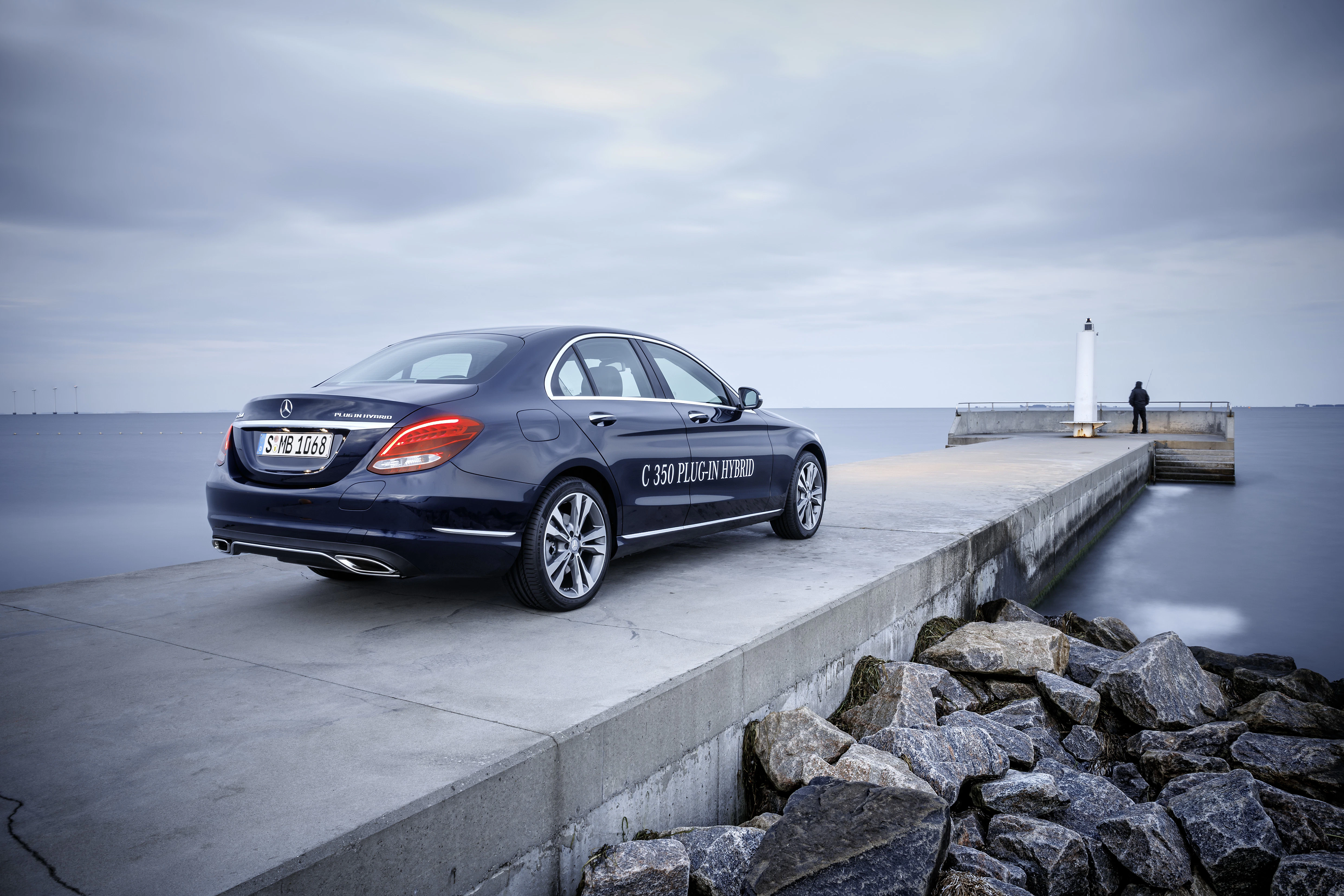 Blauer Mercedes-Benz C 350 Plug-in Hybrid auf einem Pier am Meer, ein Leuchtturm im Hintergrund unter bewölktem Himmel.