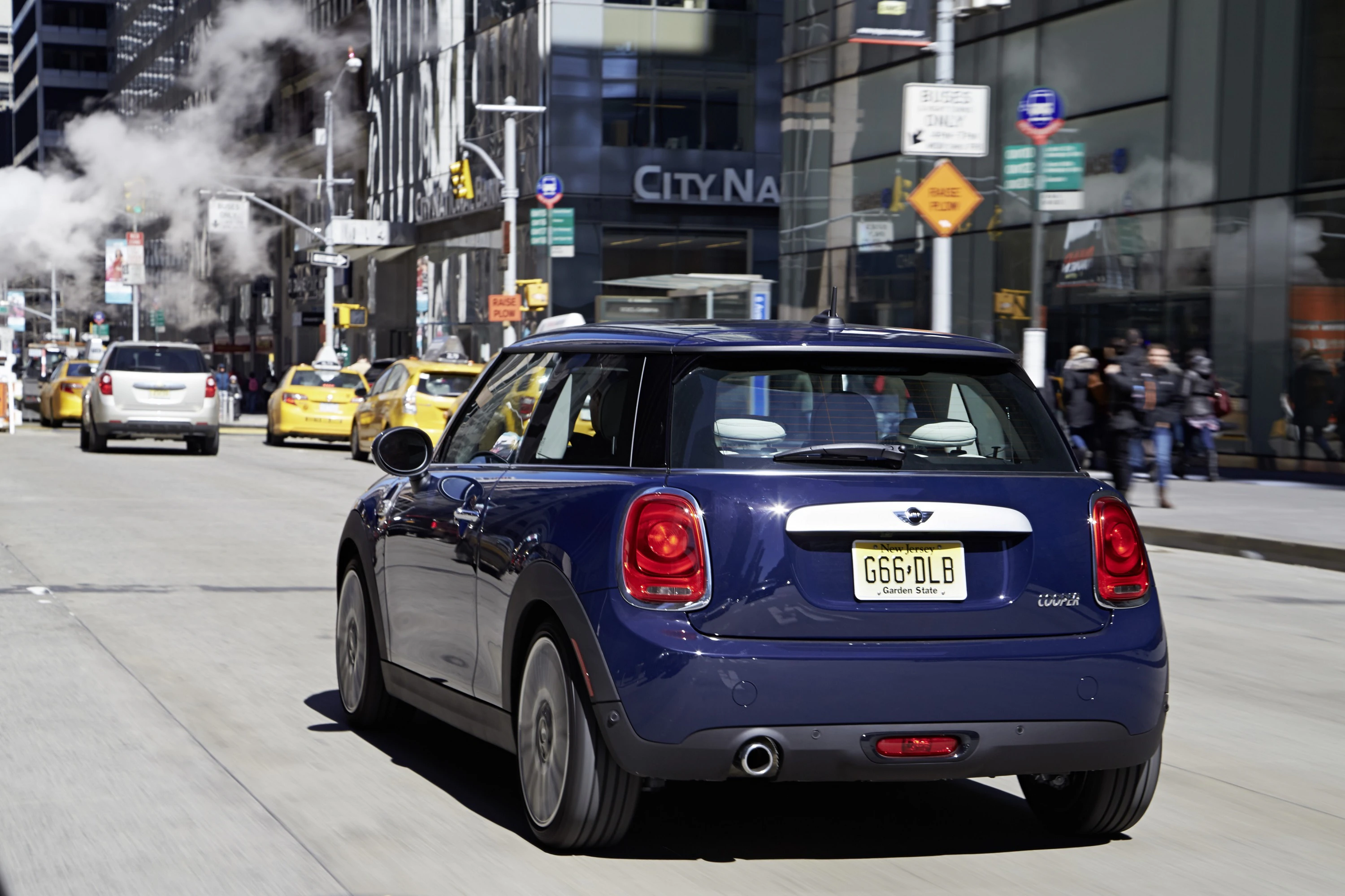 Blauer Mini Cooper fährt auf belebter Stadtstraße mit Taxis im Hintergrund. Rauch steigt aus einer Lüftung auf. Schilder und Menschen am Gehweg.