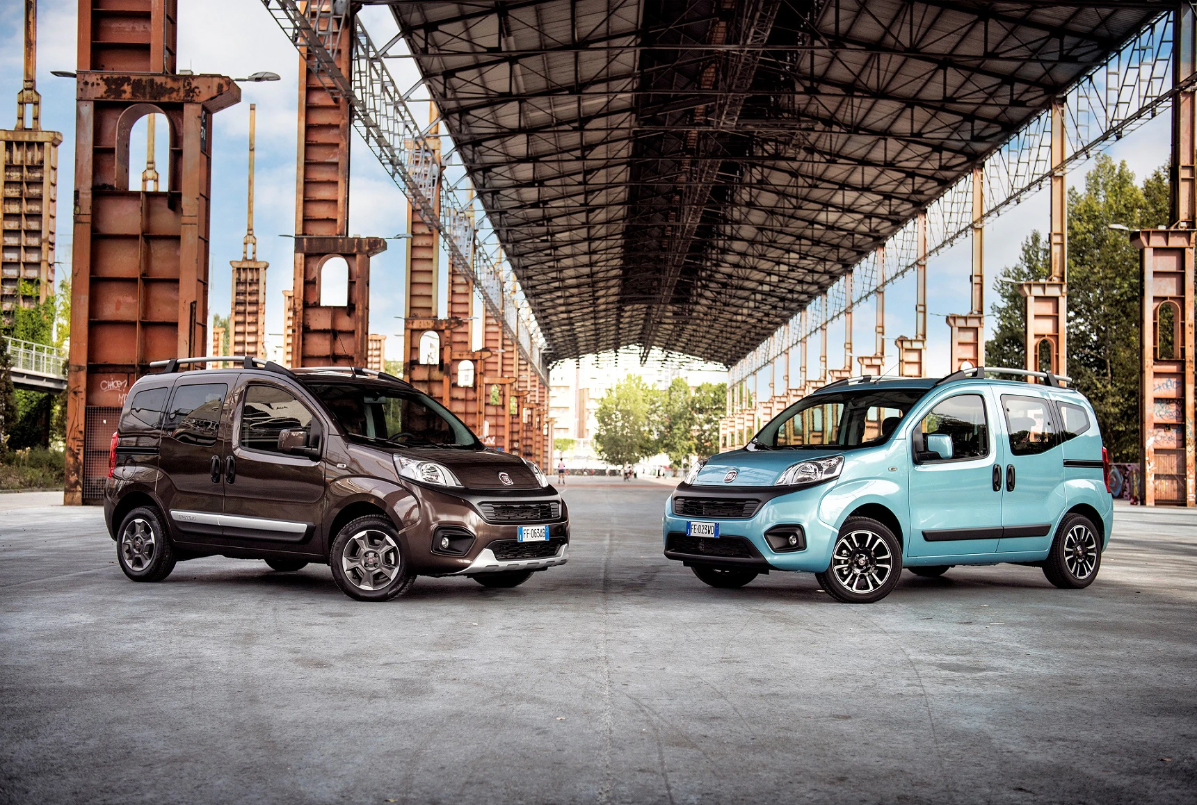 Zwei Kleintransporter stehen unter einer großen Metallkonstruktion. Links ist ein brauner und rechts ein blauer Van. Im Hintergrund sind Bäume.