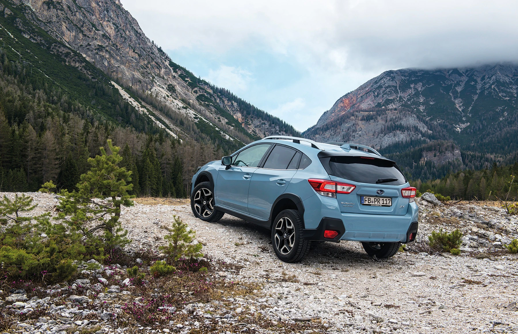 Blauer SUV auf einem schottrigen Bergpfad, umgeben von Tannen und hohen Bergen unter bewölktem Himmel.