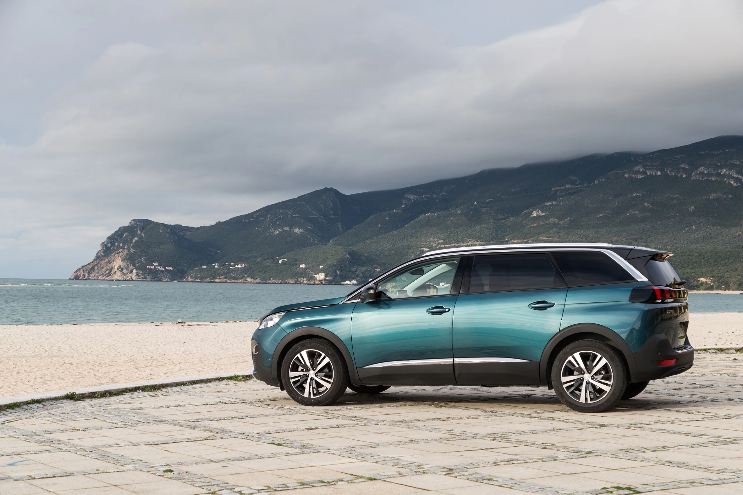 Ein blau-grüner SUV steht auf einem gepflasterten Strandweg. Im Hintergrund sind das Meer und bewaldete Berge unter einem bewölkten Himmel zu sehen.