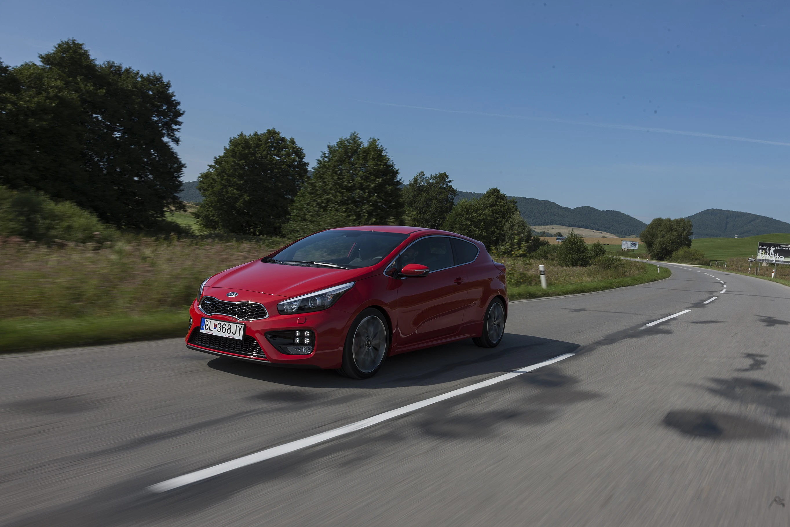 Rotes Auto fährt auf einer Landstraße durch eine grüne, hügelige Landschaft unter klarem Himmel.
