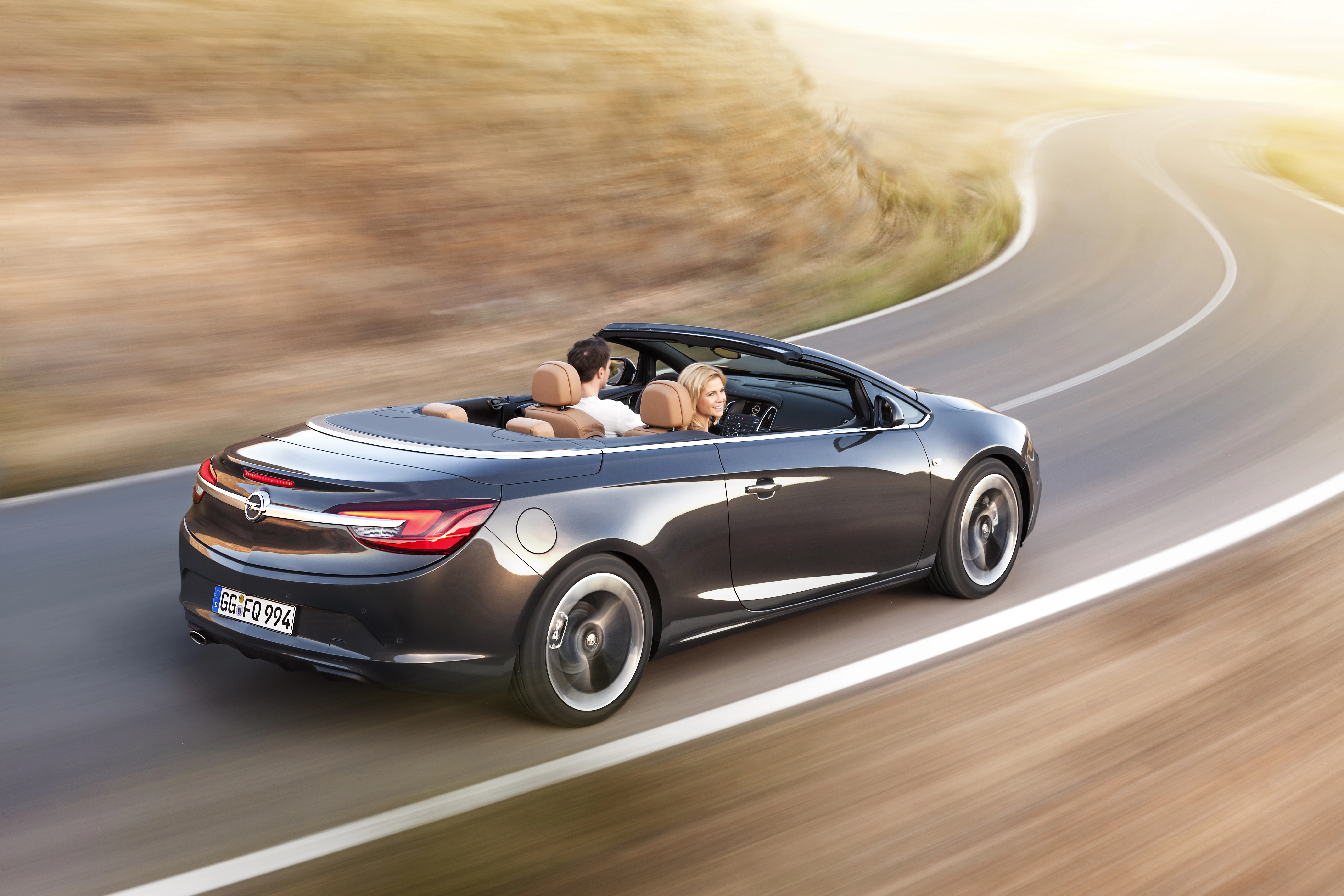 Schwarzes Cabriolet fährt mit offenem Verdeck auf kurviger Landstraße, zwei Personen an Bord, in sonniger Umgebung.