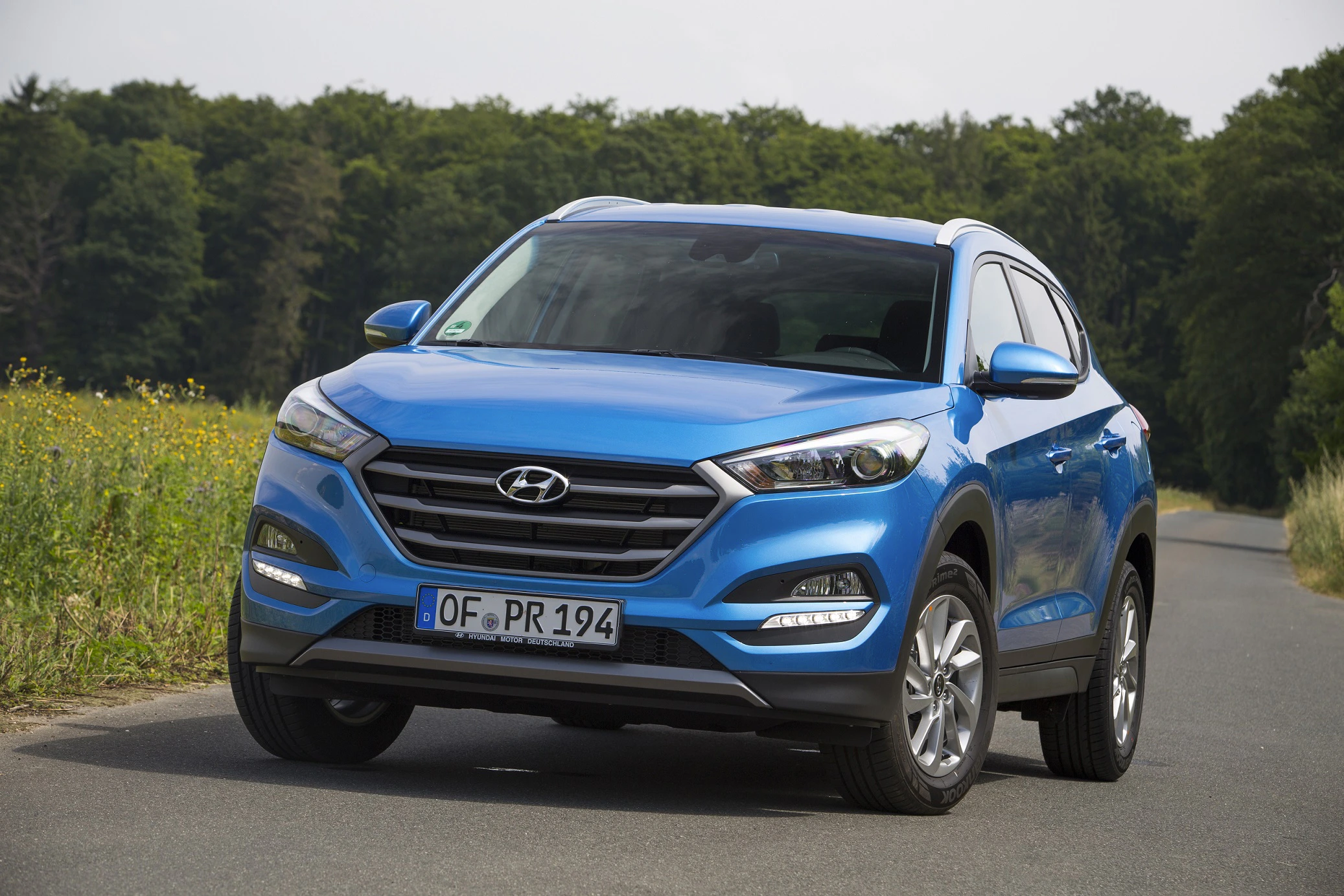 Blauer Hyundai Tucson steht auf einer Landstraße, umgeben von grüner Natur und Wald im Hintergrund.