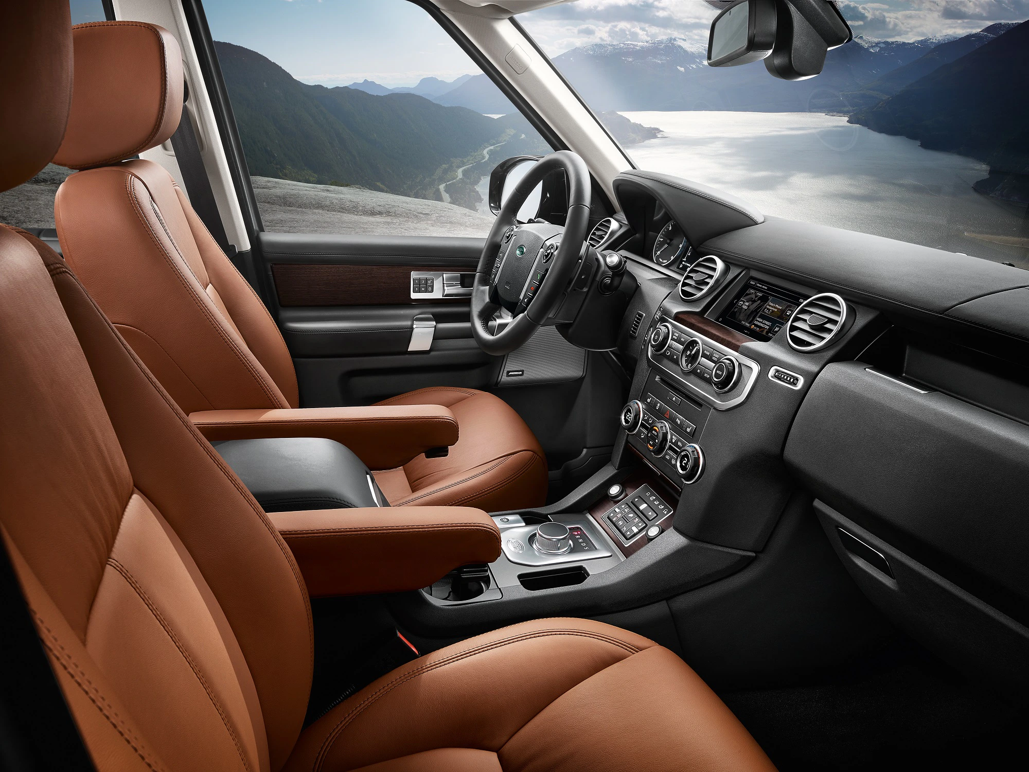 Innenraum eines luxuriösen Autos mit braunen Ledersitzen und modernem Armaturenbrett. Im Hintergrund ein malerischer Blick auf Berge und einen See.
