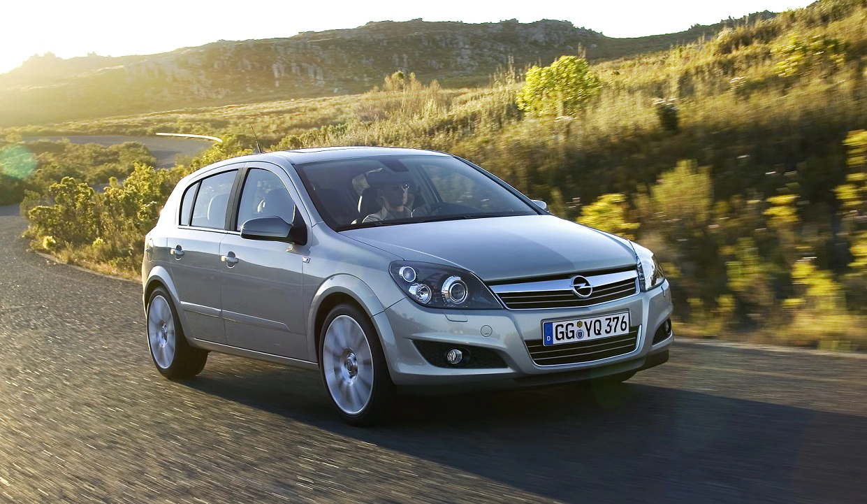 Silberner Opel Astra fährt bei Sonnenuntergang auf einer Landstraße, umgeben von Hügeln und Vegetation.