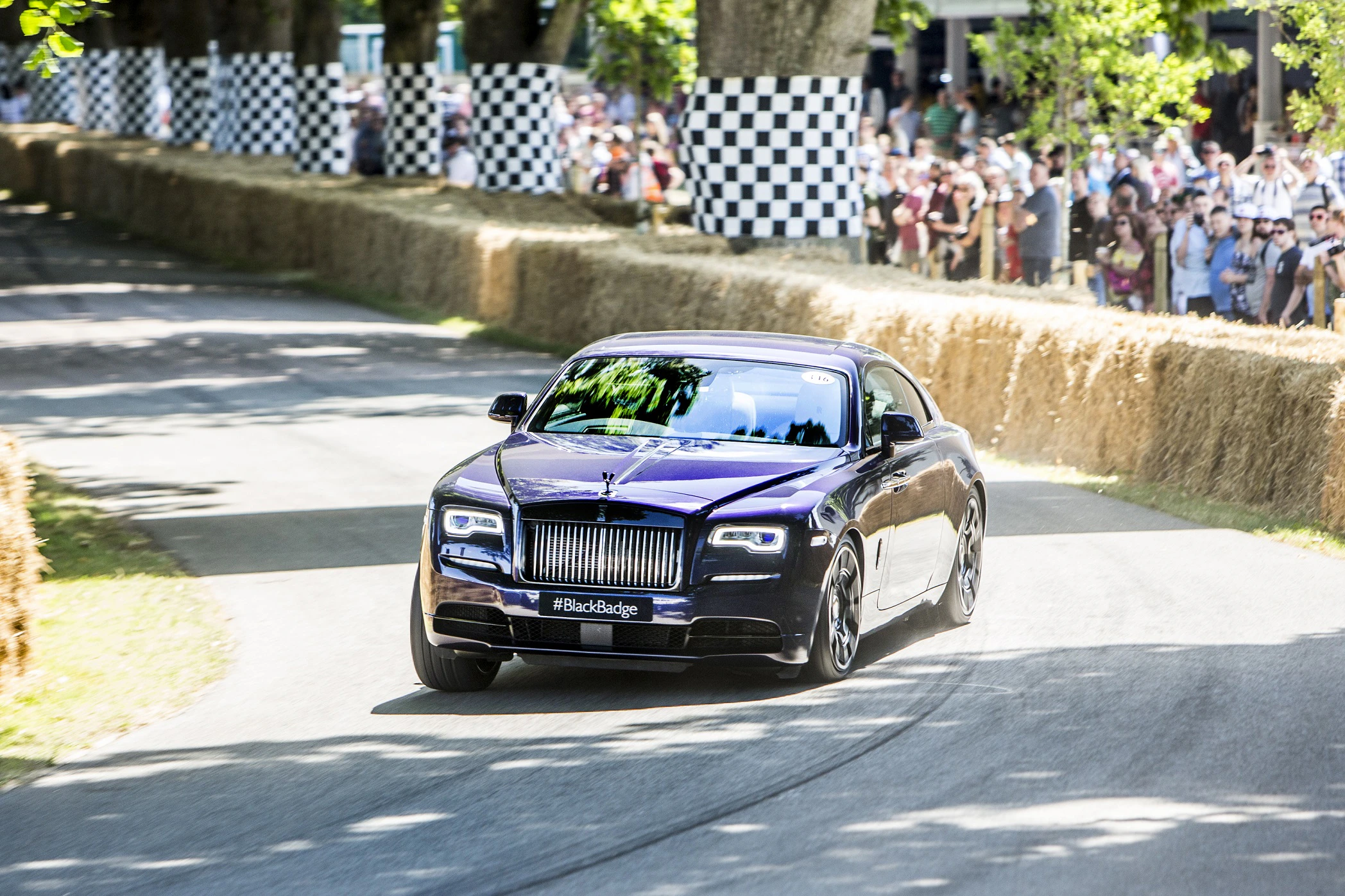 Luxuriöses, dunkelviolettes Auto fährt eine kurvige Strecke bei einem Event, umgeben von Zuschauern und Strohballen.