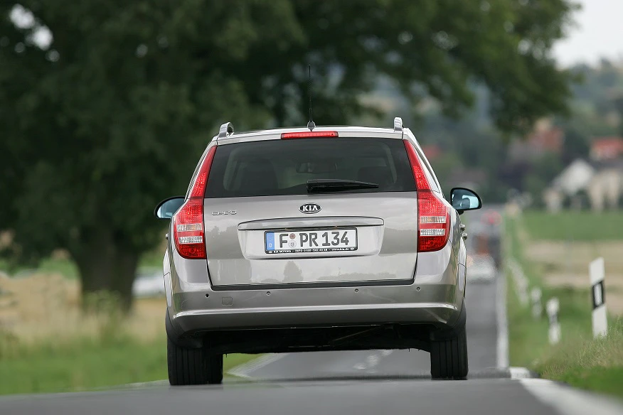 Silberner Kia cee’d Kombi fährt auf schmaler Landstraße, umgeben von Bäumen und einem Feld. Das Auto ist von hinten zu sehen.