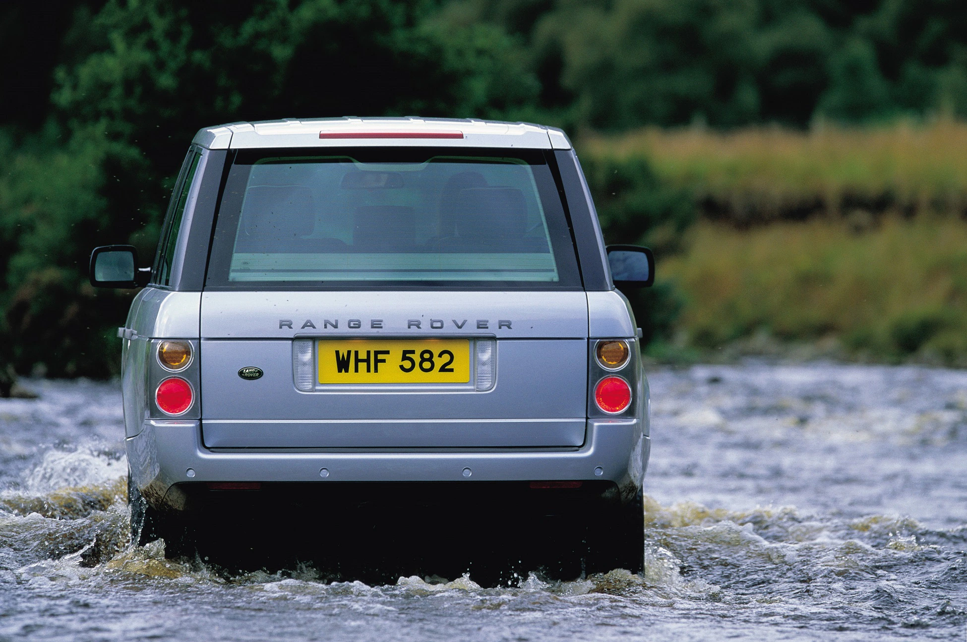 Ein silberner Range Rover fährt durch ein flaches Gewässer in einer natürlichen Umgebung.