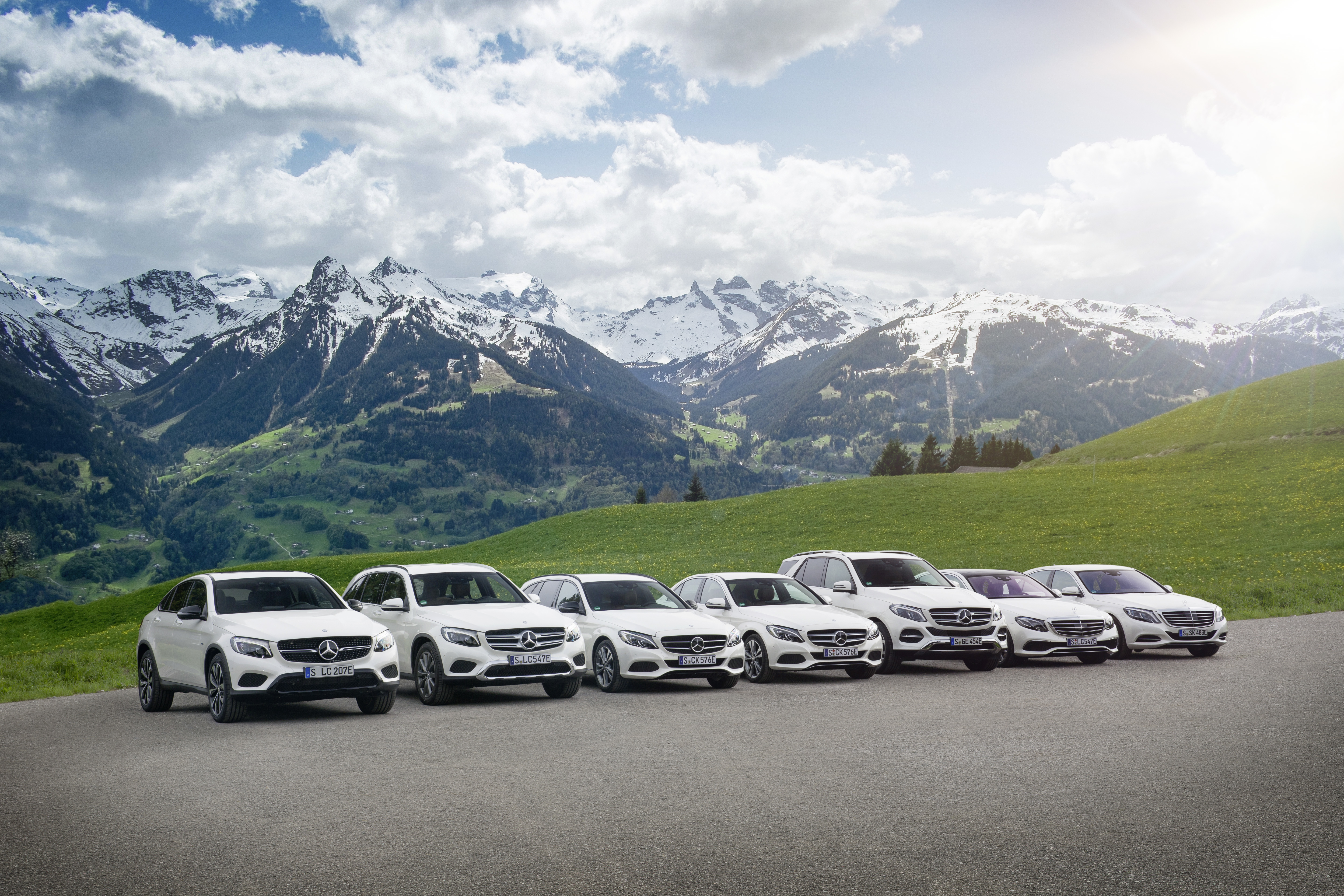 Eine Reihe weißer Mercedes-Benz-Modelle steht vor einer malerischen Alpenlandschaft mit grünen Hügeln und schneebedeckten Bergen unter blauem Himmel.