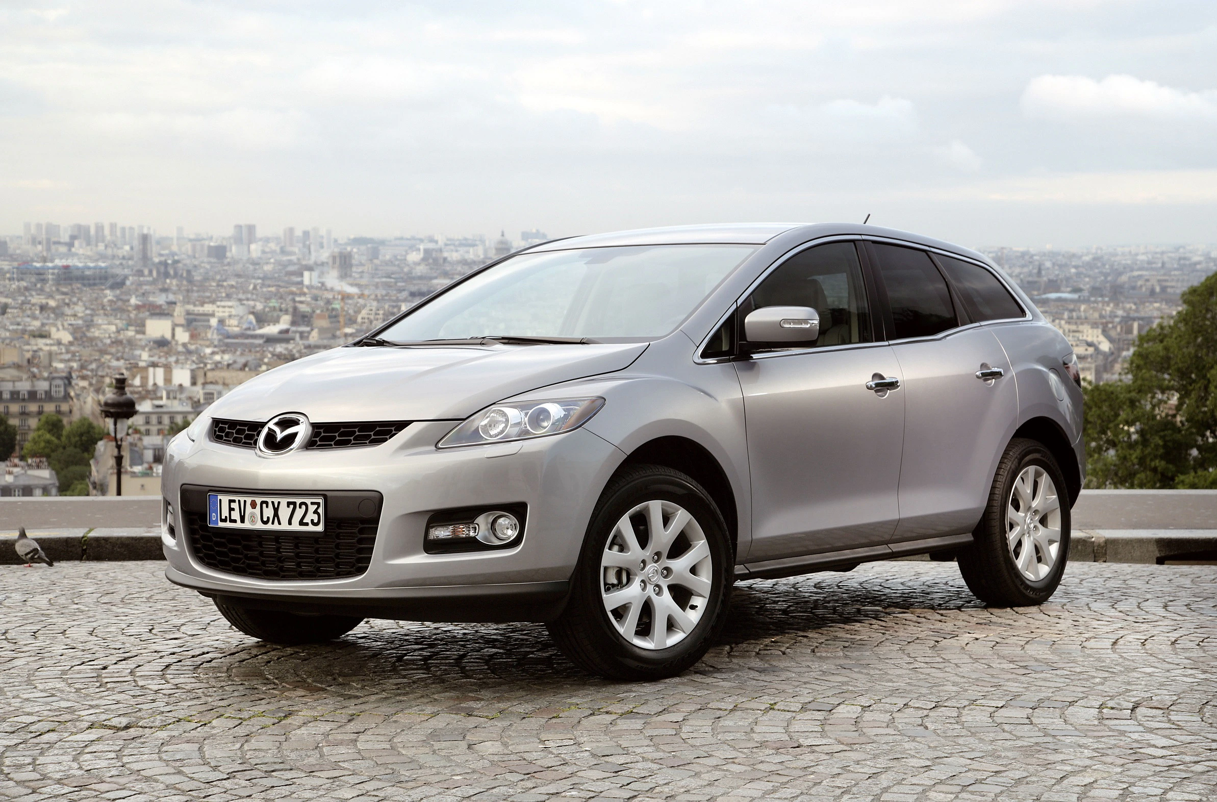 Silberner Mazda CX-7 steht auf einem Kopfsteinpflaster mit Stadtpanorama im Hintergrund an einem bewölkten Tag.