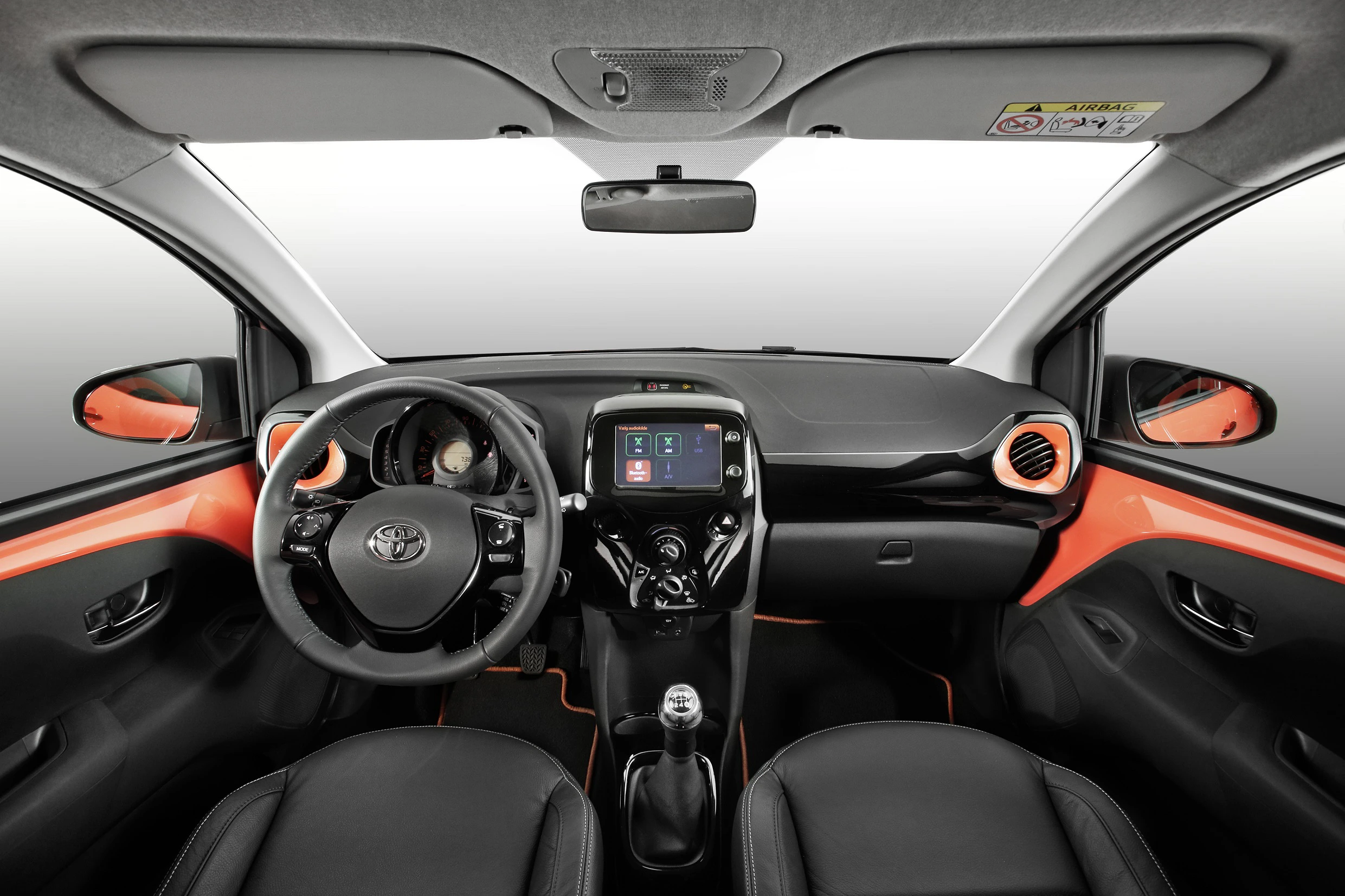 Innenraum eines modernen Autos mit orangefarbenen Akzenten, Lenkrad links, zentralem Bildschirm und zwei schwarzen Sitzen.