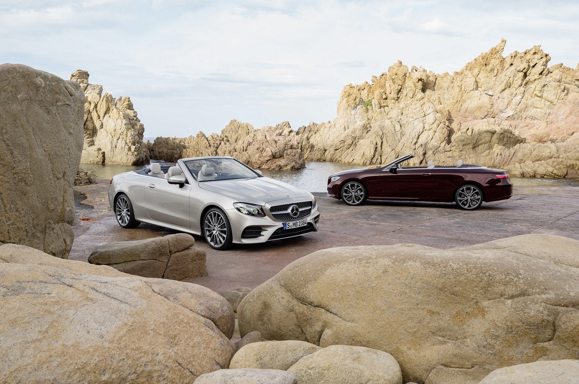 Zwei Cabriolets in einer felsigen Küstenlandschaft, ein silbernes und ein dunkelrotes Auto, mit geöffnetem Verdeck auf einem asphaltierten Weg.