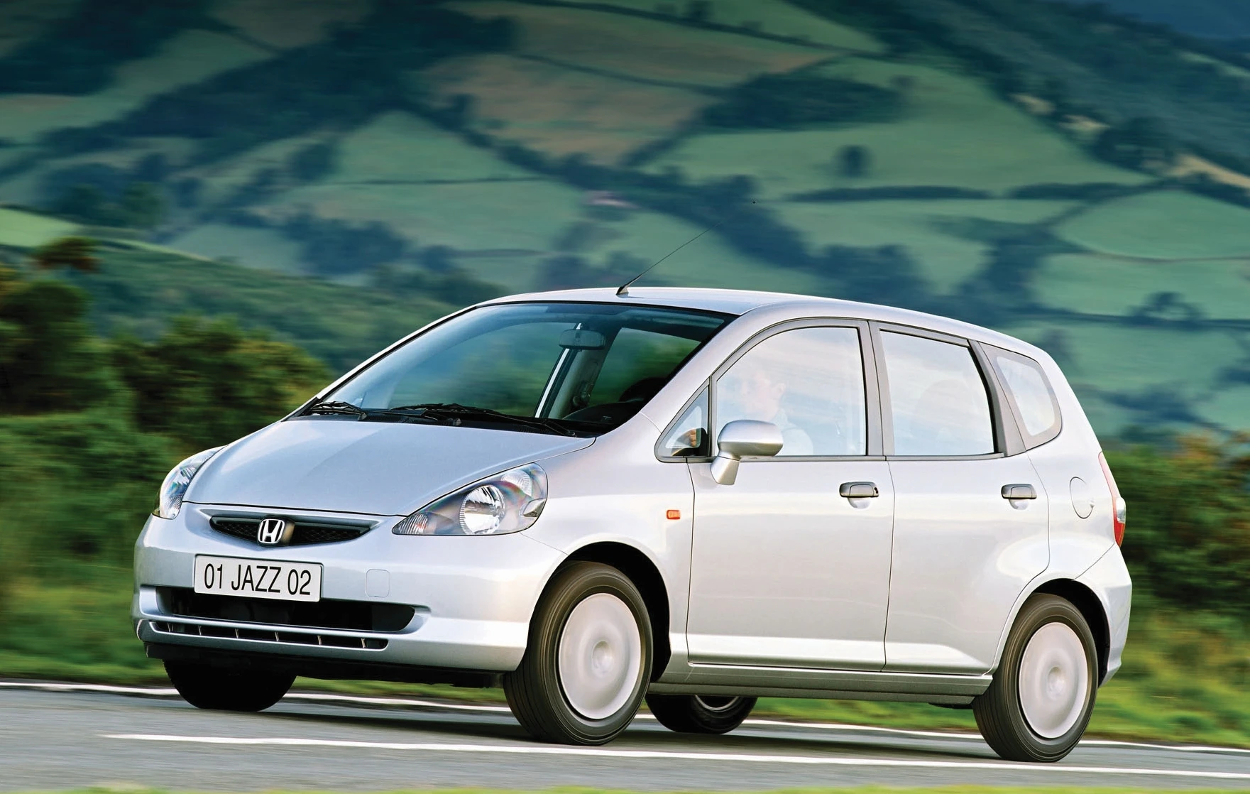 Silberner Honda Jazz fährt auf einer Landstraße, im Hintergrund grüne Felder und Hügel.