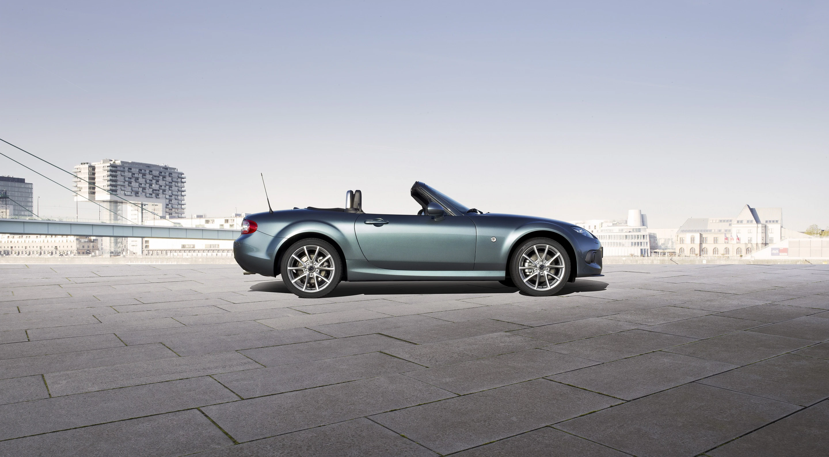 Ein blaues Cabrio-Pkw steht auf einem großen Pflasterplatz, mit einer modernen Stadtlandschaft im Hintergrund unter klarem Himmel.